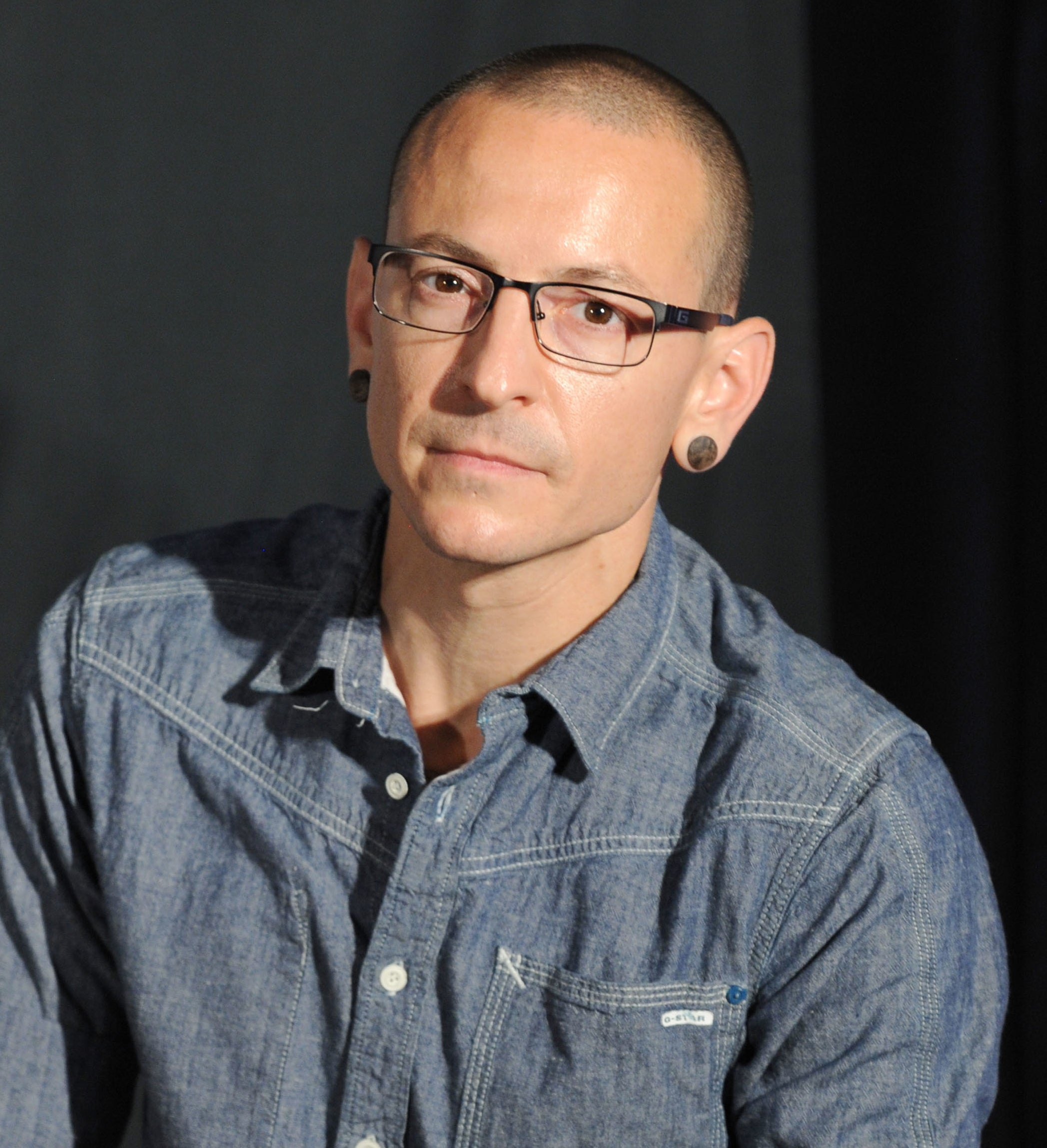 Chester Bennington with short hair and glasses, wearing a rolled-up denim shirt and showing arm tattoos, stands against a plain background