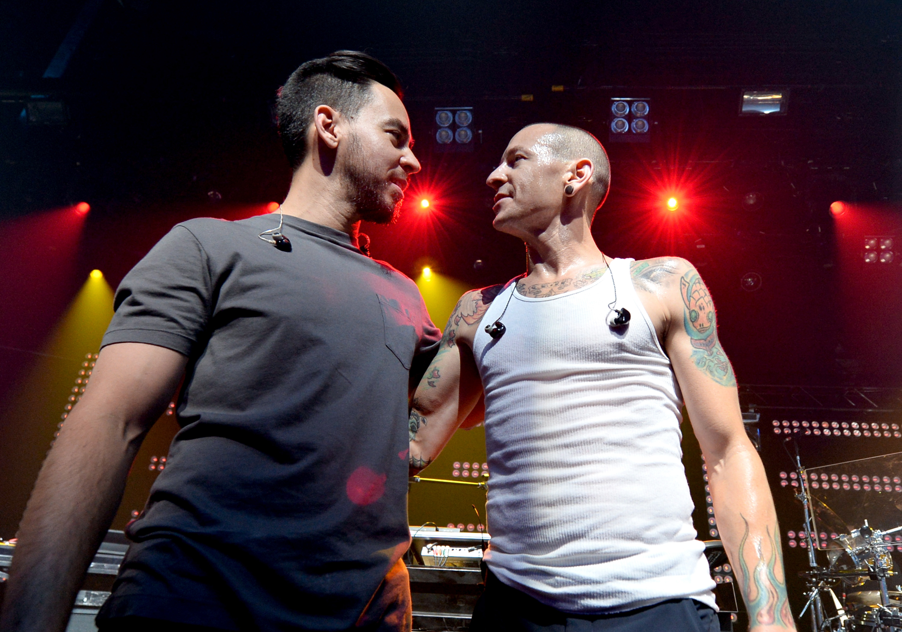 Mike Shinoda and Chester Bennington on stage in a close embrace, both wearing casual clothing and visible earpieces, under red stage lights