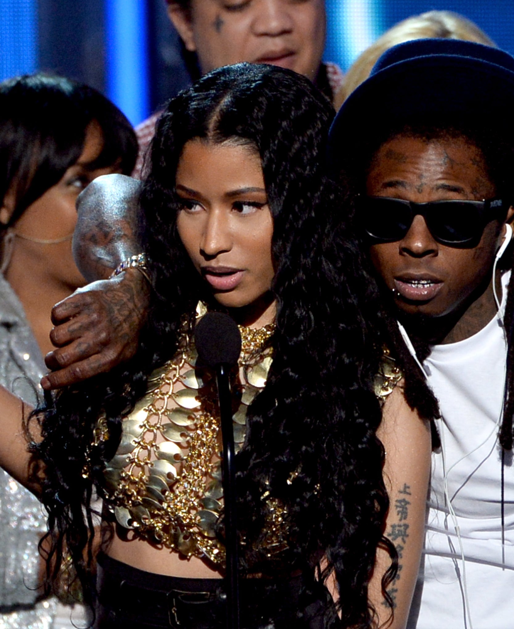 Nicki Minaj and Lil Wayne on stage at an event with others in the background. Nicki is holding a microphone, and Lil Wayne has his arm around her