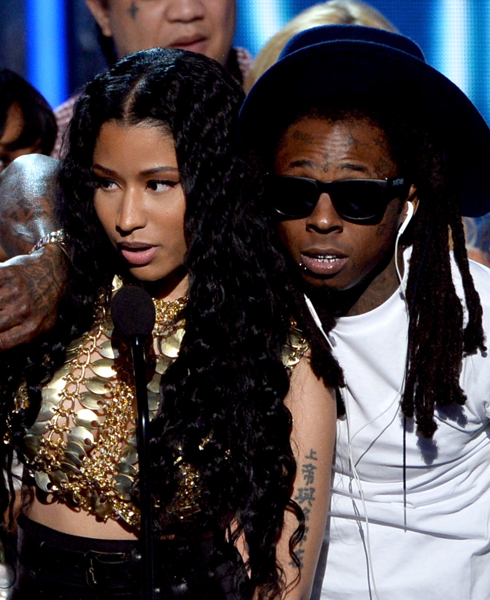 Nicki Minaj and Lil Wayne on stage at an event with others in the background. Nicki is holding a microphone, and Lil Wayne has his arm around her