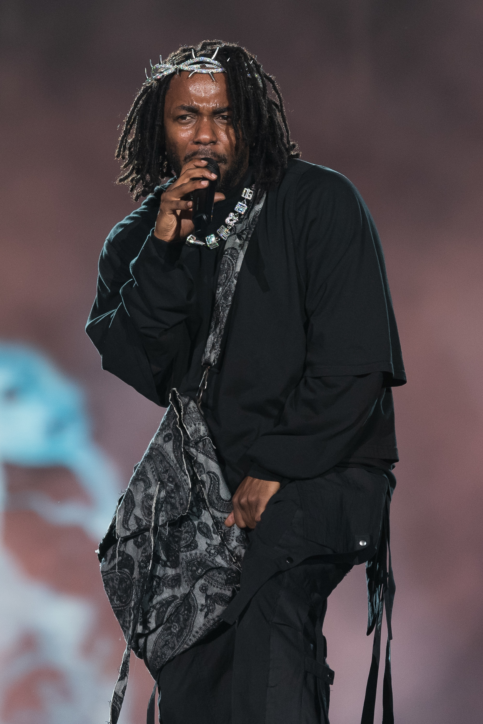 Kendrick Lamar performs on stage, holding a mic. He wears a loose outfit with intricate patterns and beaded accessories