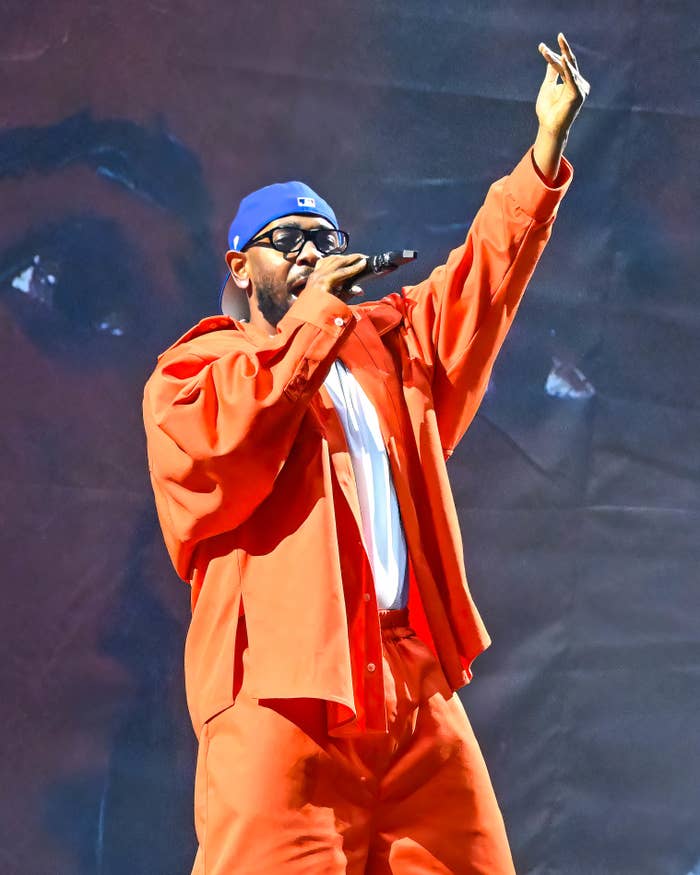 Kendrick Lamar performs on stage, wearing a cap, glasses, and an bright outfit, raising one hand while holding a microphone with the other