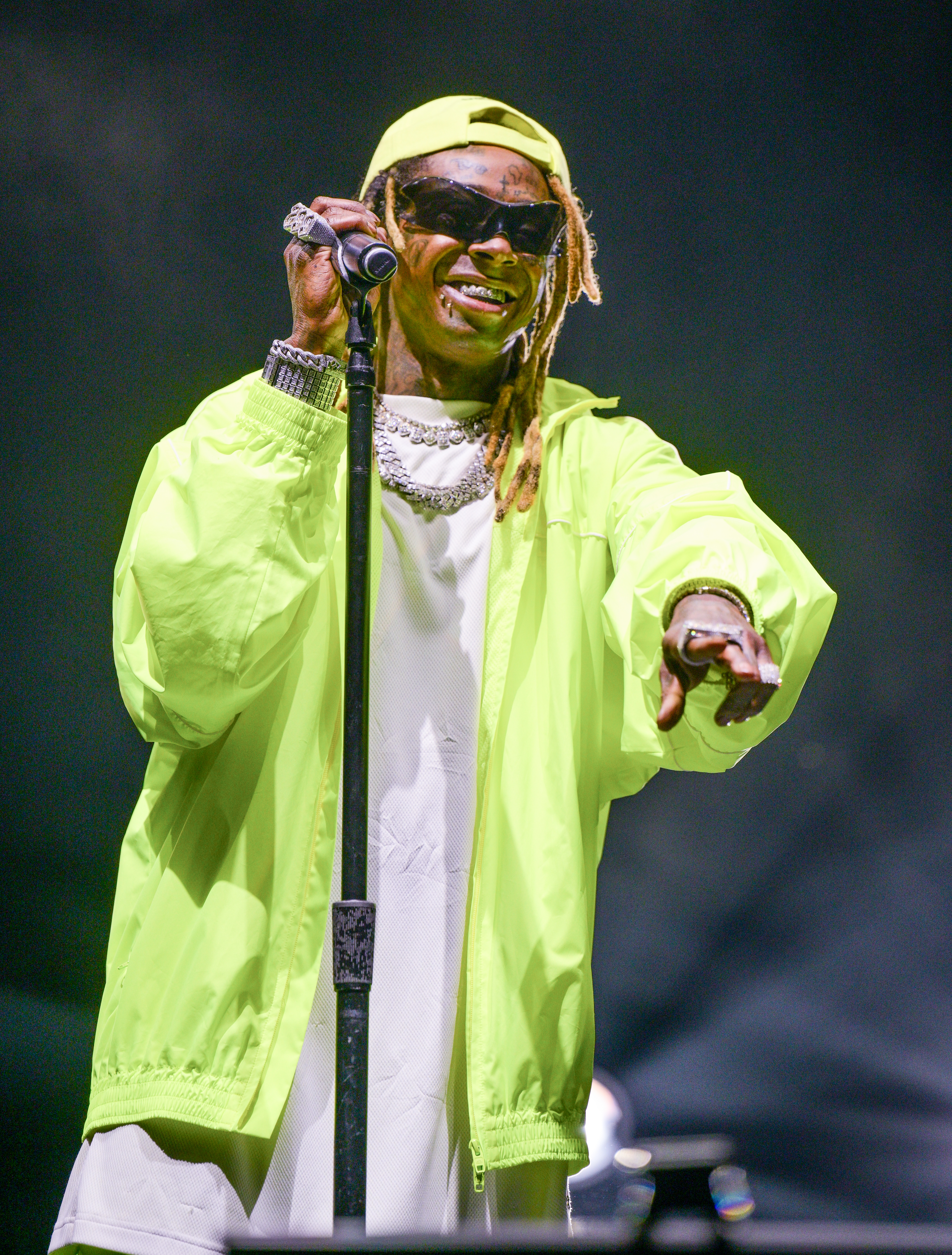 Lil Wayne performs on stage wearing a neon jacket, shirt, sunglasses, and a cap. He holds a microphone and points towards the audience