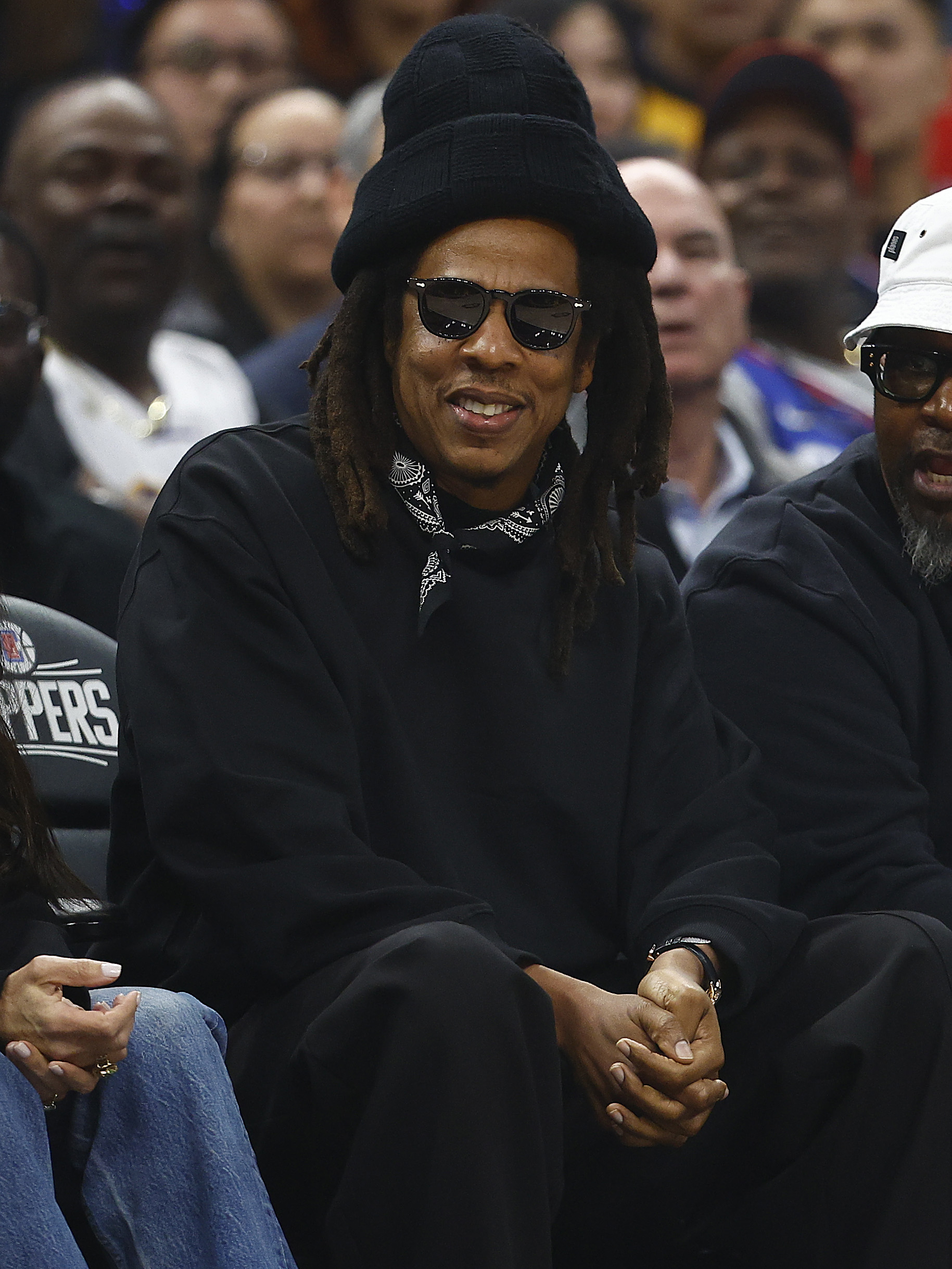 Jay-Z sits courtside at a basketball game, wearing a hat, sunglasses, a sweatshirt, and a bandana around his neck