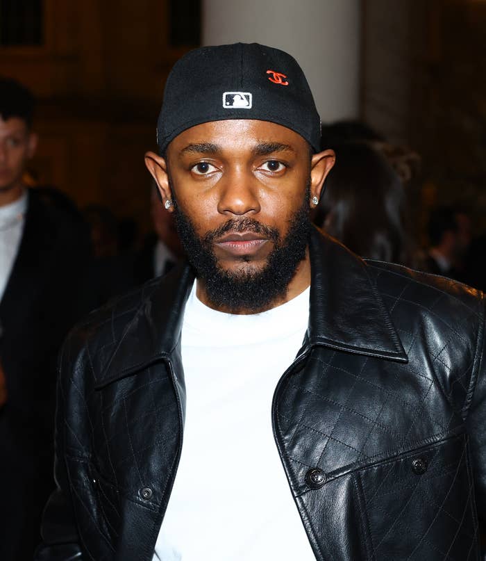 Kendrick Lamar wearing a leather jacket and a cap, posing at an event with other attendees in the background