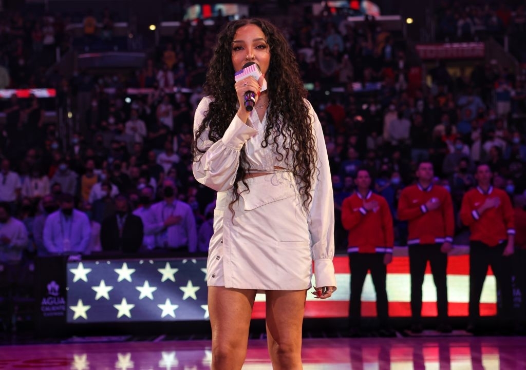 Tinashe sings into a microphone on a basketball court, dressed in a stylish outfit, with a crowd and an American flag in the background
