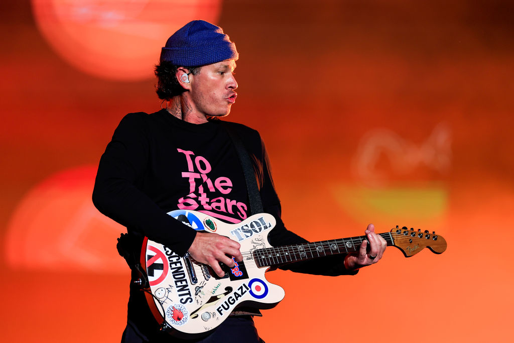 Tom DeLonge playing guitar on stage, wearing a &quot;To The Stars&quot; shirt and a beanie. His guitar is covered with various stickers