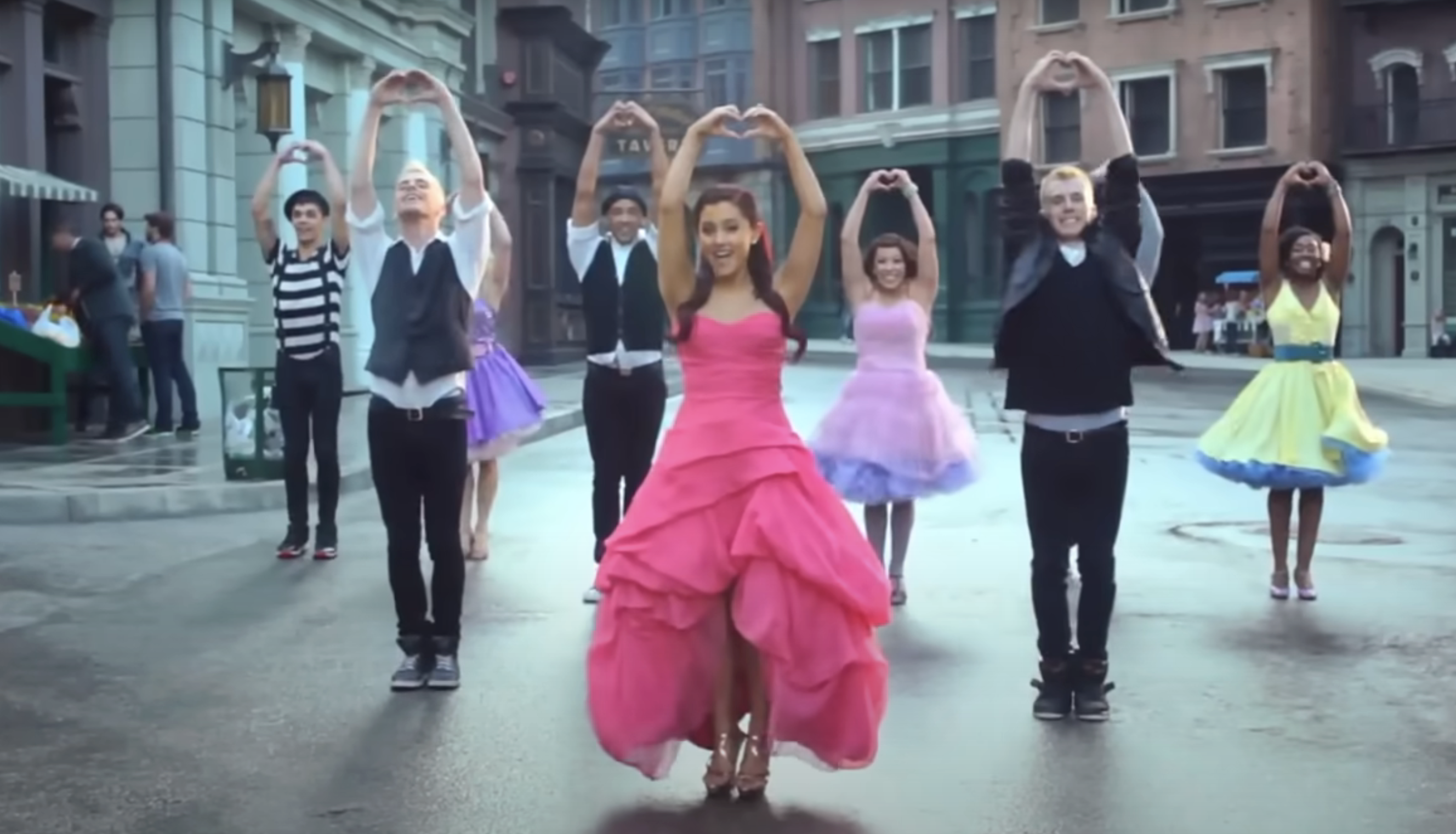 Ariana Grande performs with dancers in heart pose on a street set. She's in a ruffled dress; dancers wear coordinated outfits