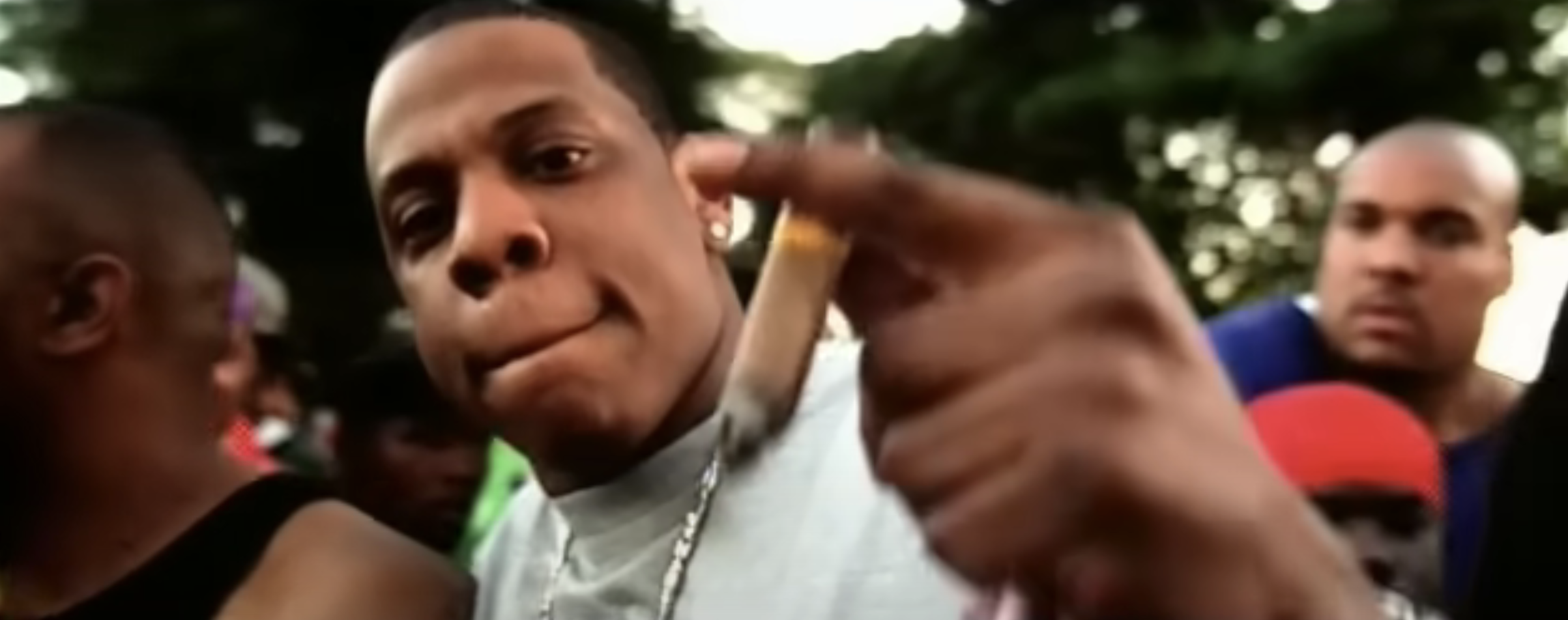 Jay-Z is holding a cigar and pointing towards the camera in a crowded outdoor setting