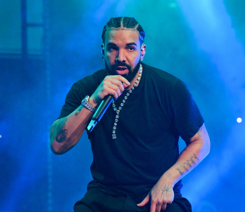 Drake performs on stage, holding a microphone with one hand and kneeling, wearing a dark T-shirt, and multiple silver chains, against a vibrant background