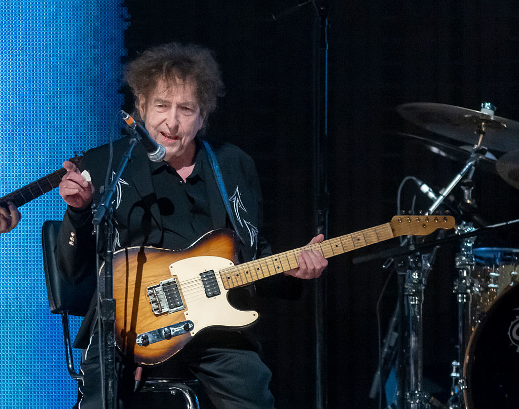 Bob Dylan playing guitar on stage in a live performance, wearing a dark outfit
