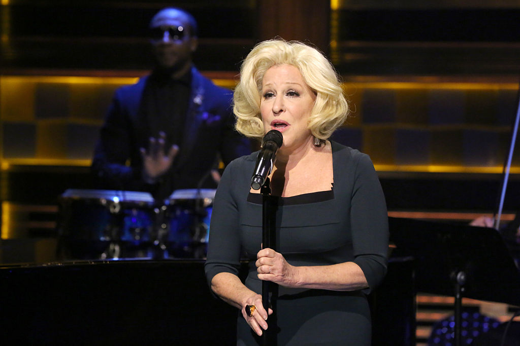 Bette Midler sings into a microphone on stage during a musical performance, accompanied by a band