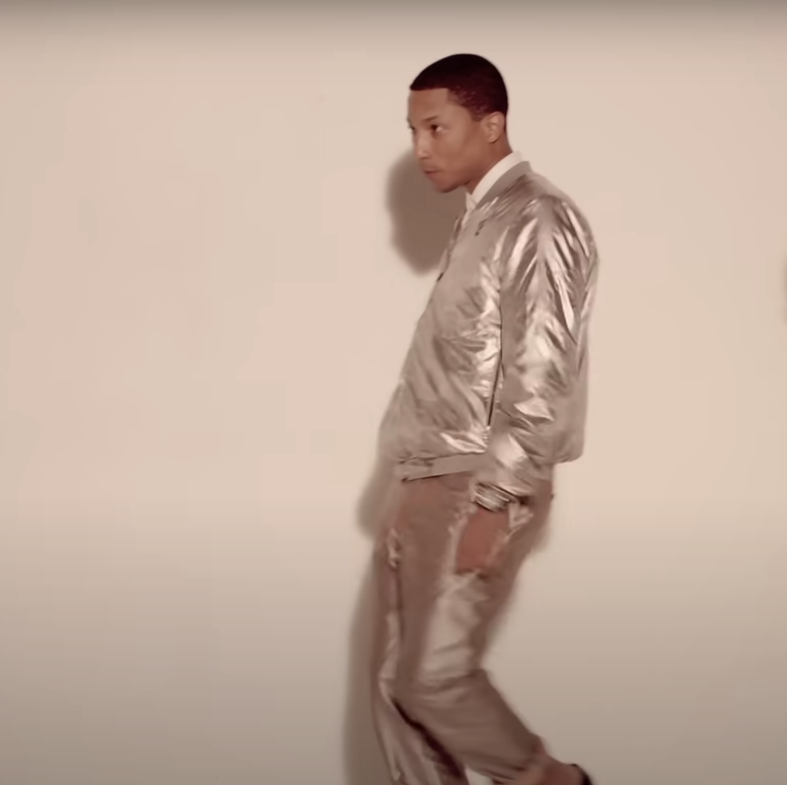 Pharrell Williams wearing a shiny, metallic outfit, walking against a plain background