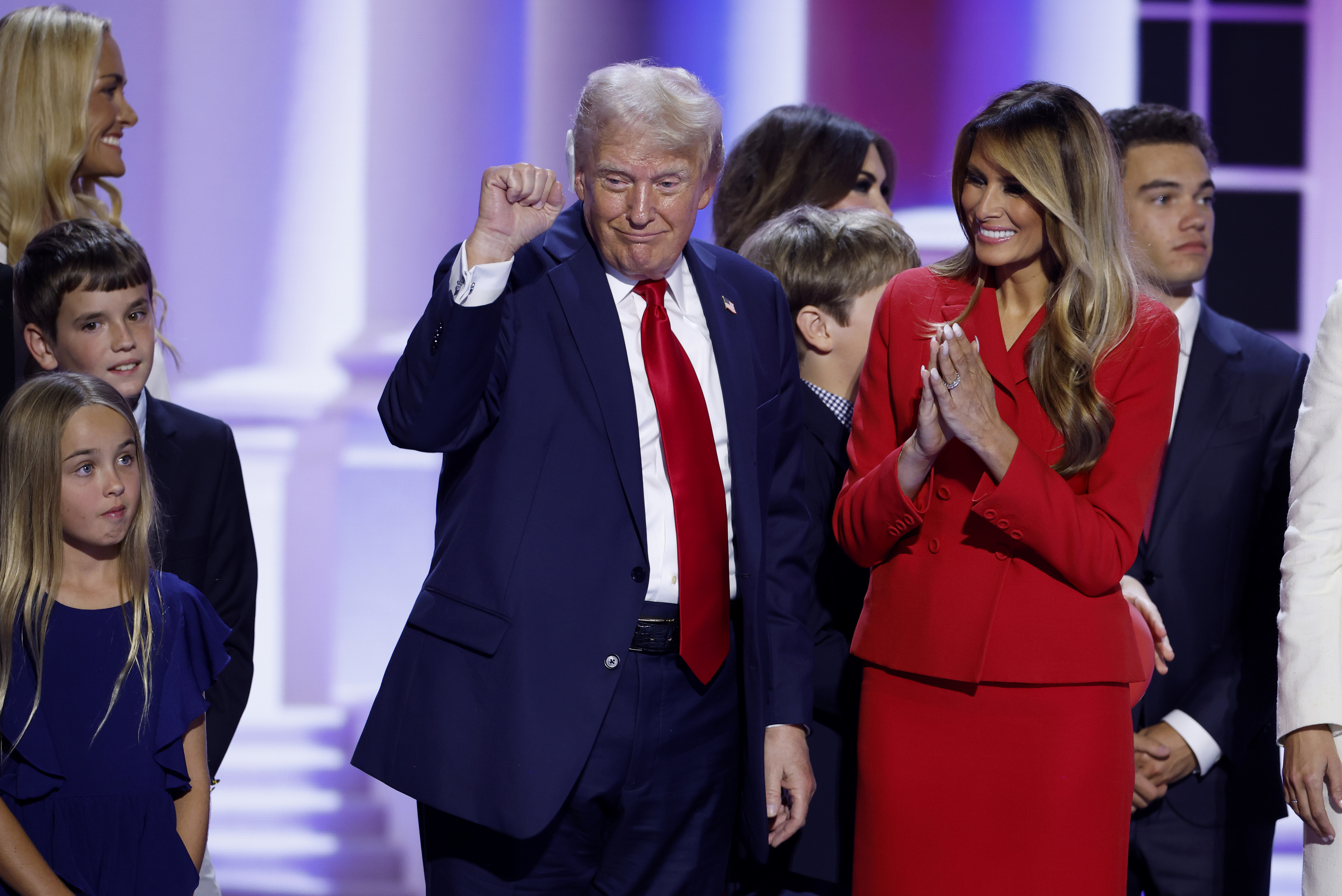 Donald Trump, Melania Trump, and others including children, on stage at an event. Trump raises his fist while Melania applauds