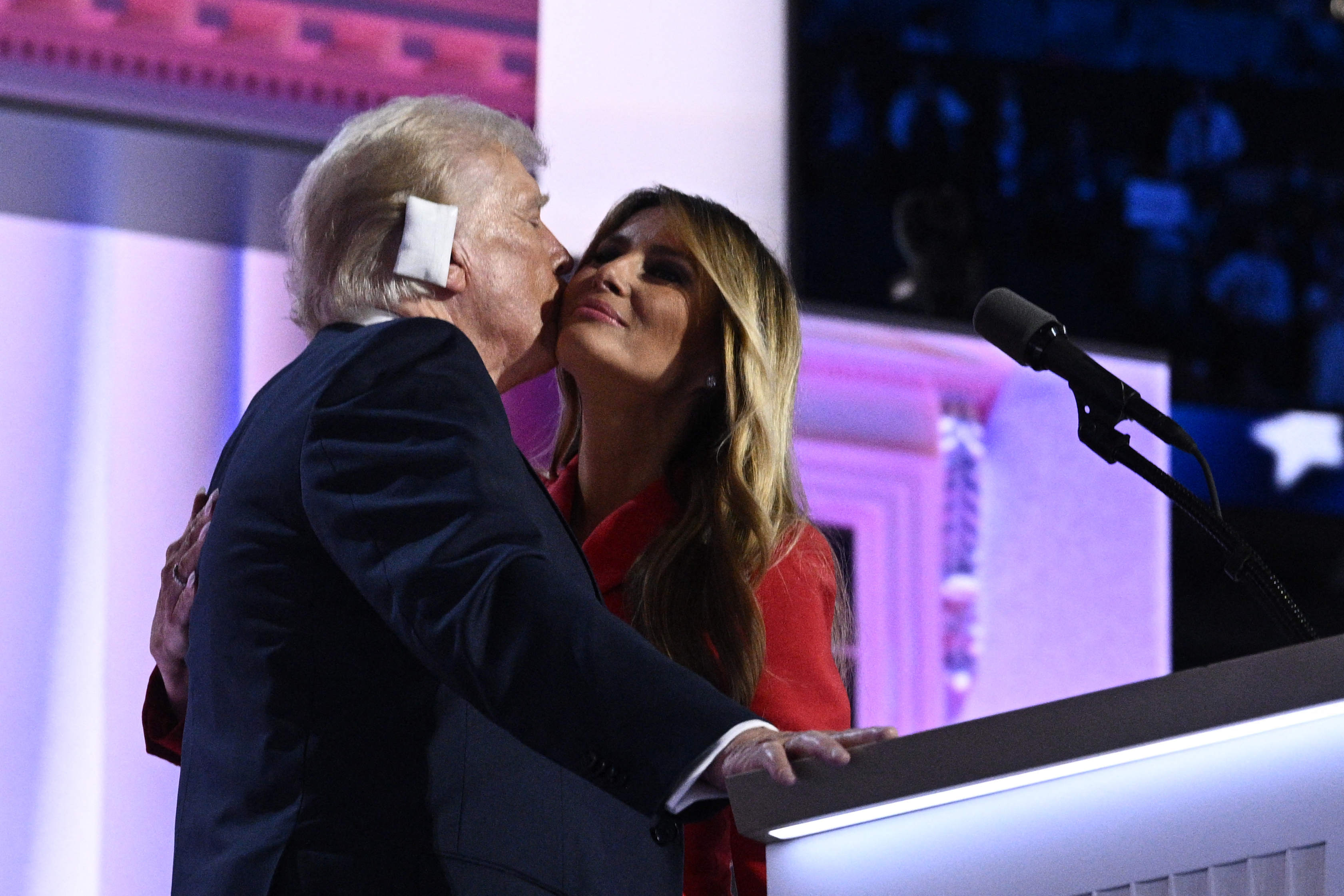 Donald Trump kisses Melania Trump on a stage with a microphone. Melania wears a formal outfit
