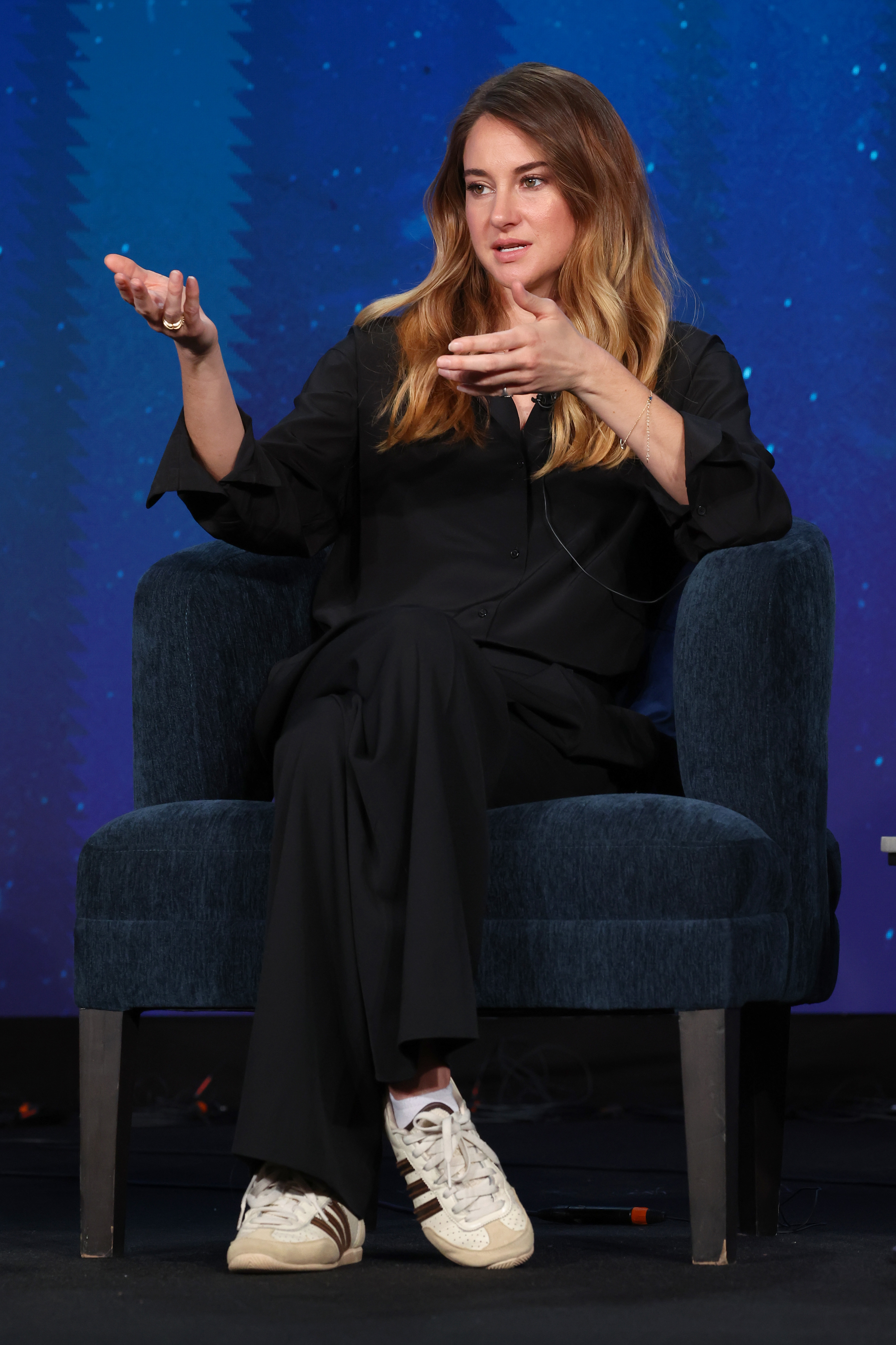 Shailene Woodley speaking onstage, seated in a chair, wearing a loose-fitting dark outfit and white sneakers, gesturing with one hand