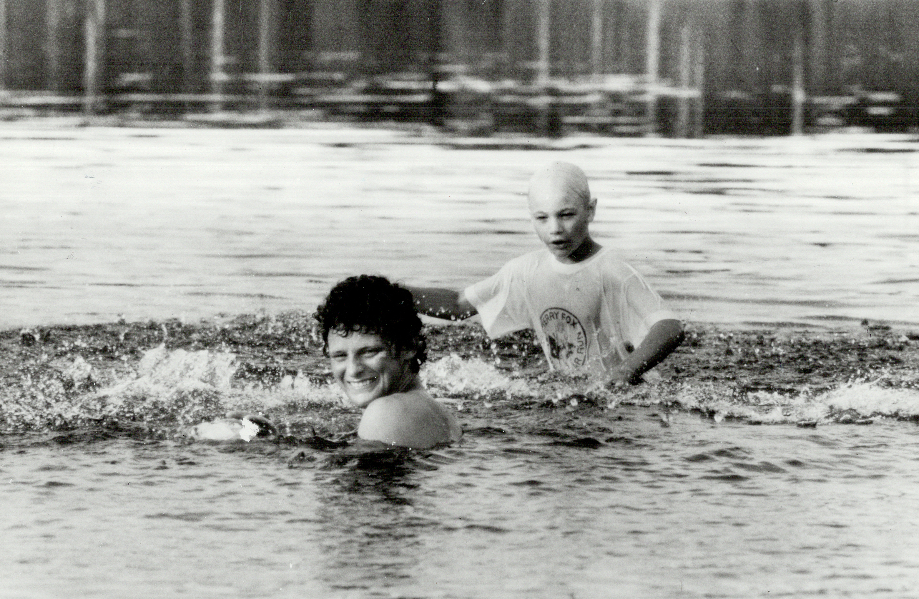 Two individuals swimming in a lake, captured in a candid moment. The person with short hair is submerged up to their shoulders and smiling, the other wears a swim cap and a t-shirt
