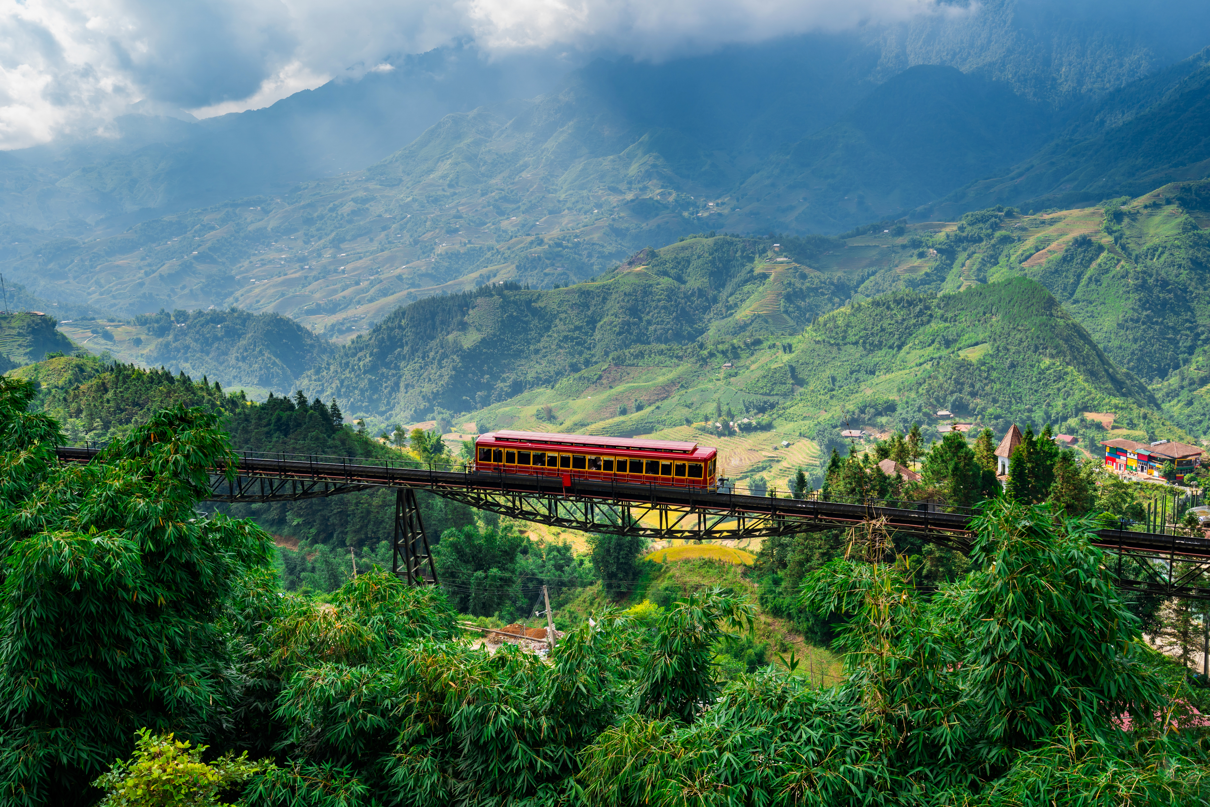 A red train crosses a high bridge amidst lush green mountains and valleys. The sky is partly cloudy, adding to the scenic beauty