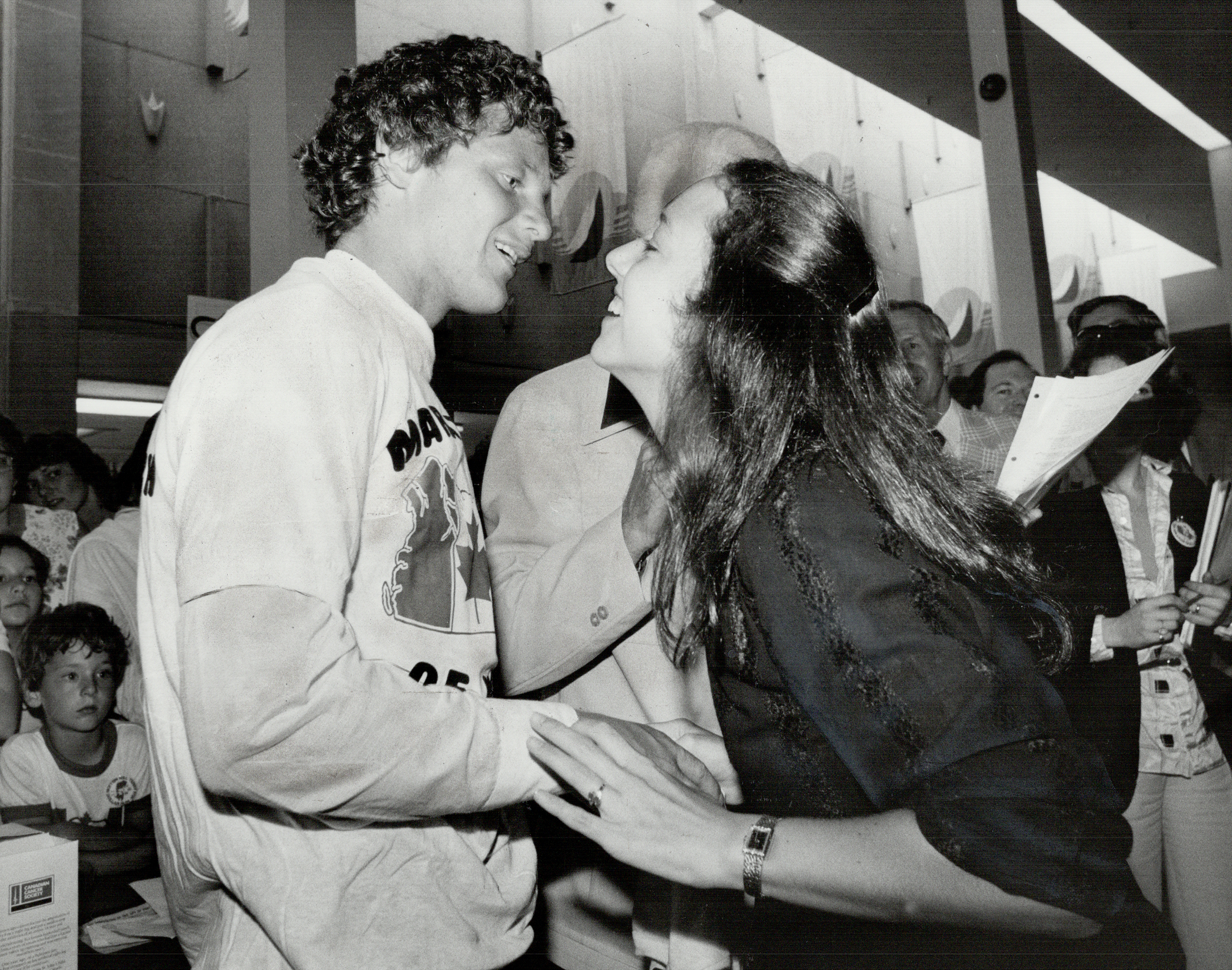 Terry Fox, wearing a Marathon of Hope shirt, is greeted warmly by a woman in a busy public setting. Fans and supporters are seen in the background
