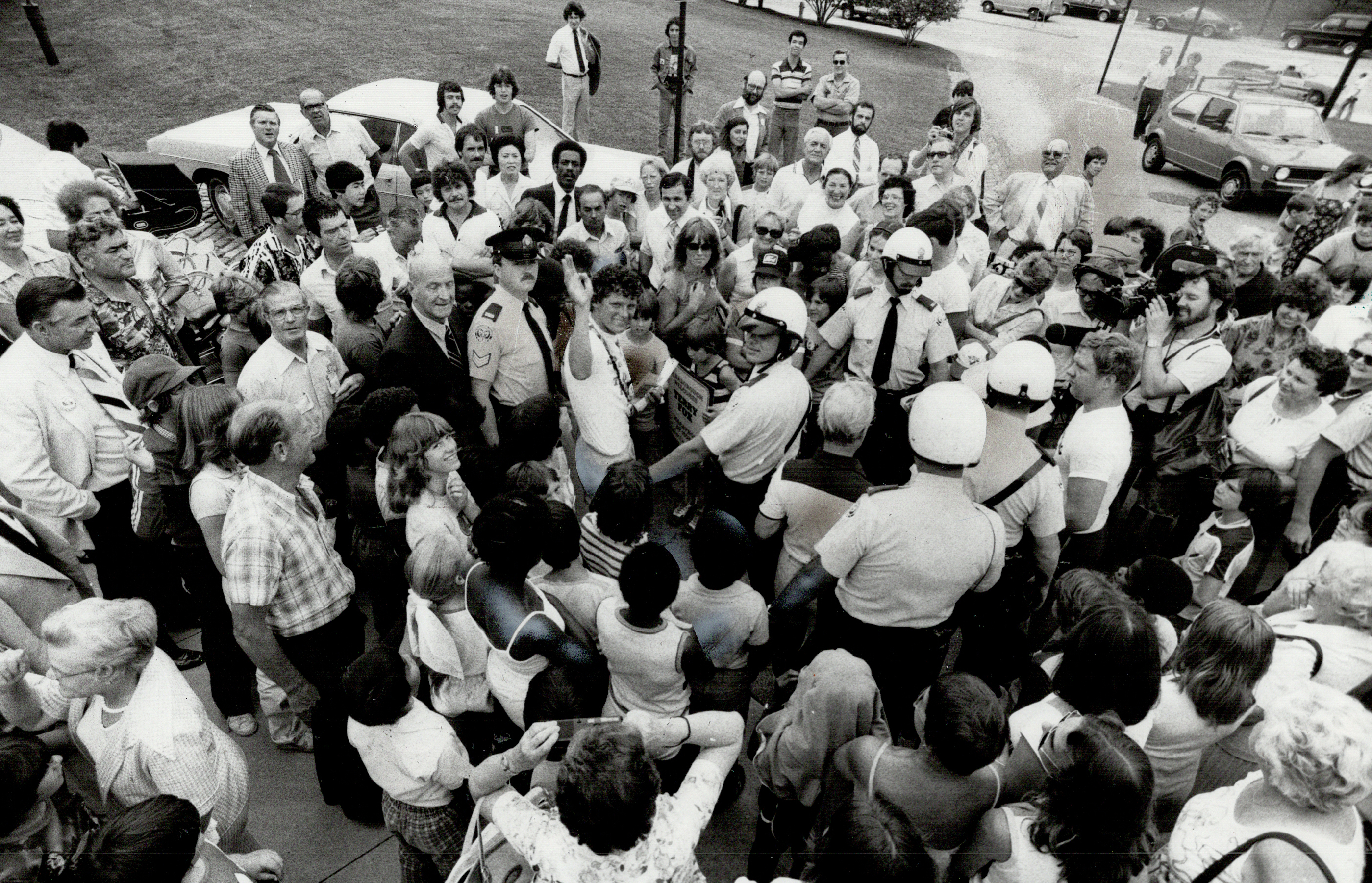 A large crowd gathers around a group of police officers and community leaders during a street event. Bystanders of various ages observe the scene keenly
