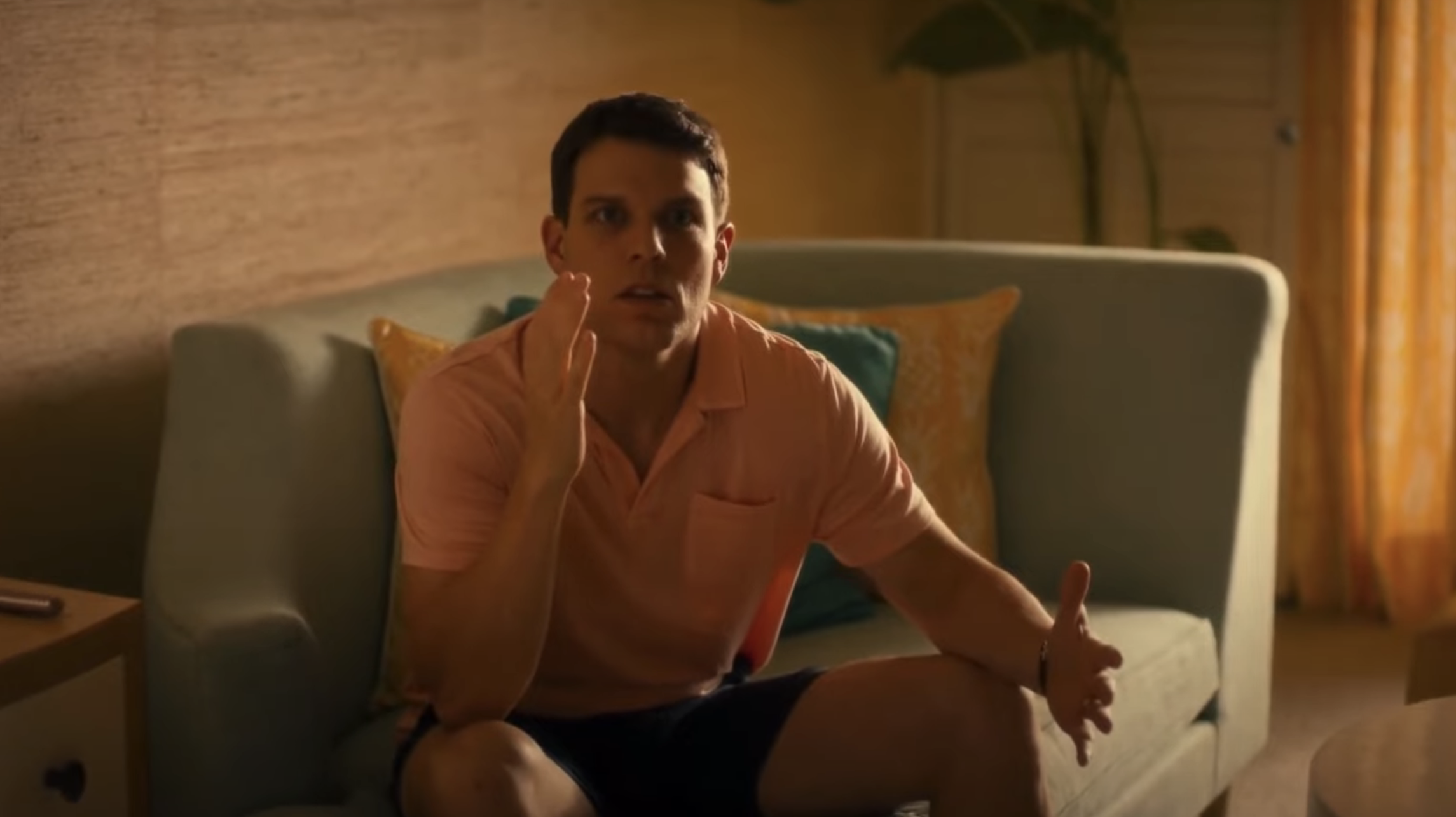 A man wearing a casual polo shirt and shorts sits on a couch, gesturing with his hands and looking focused