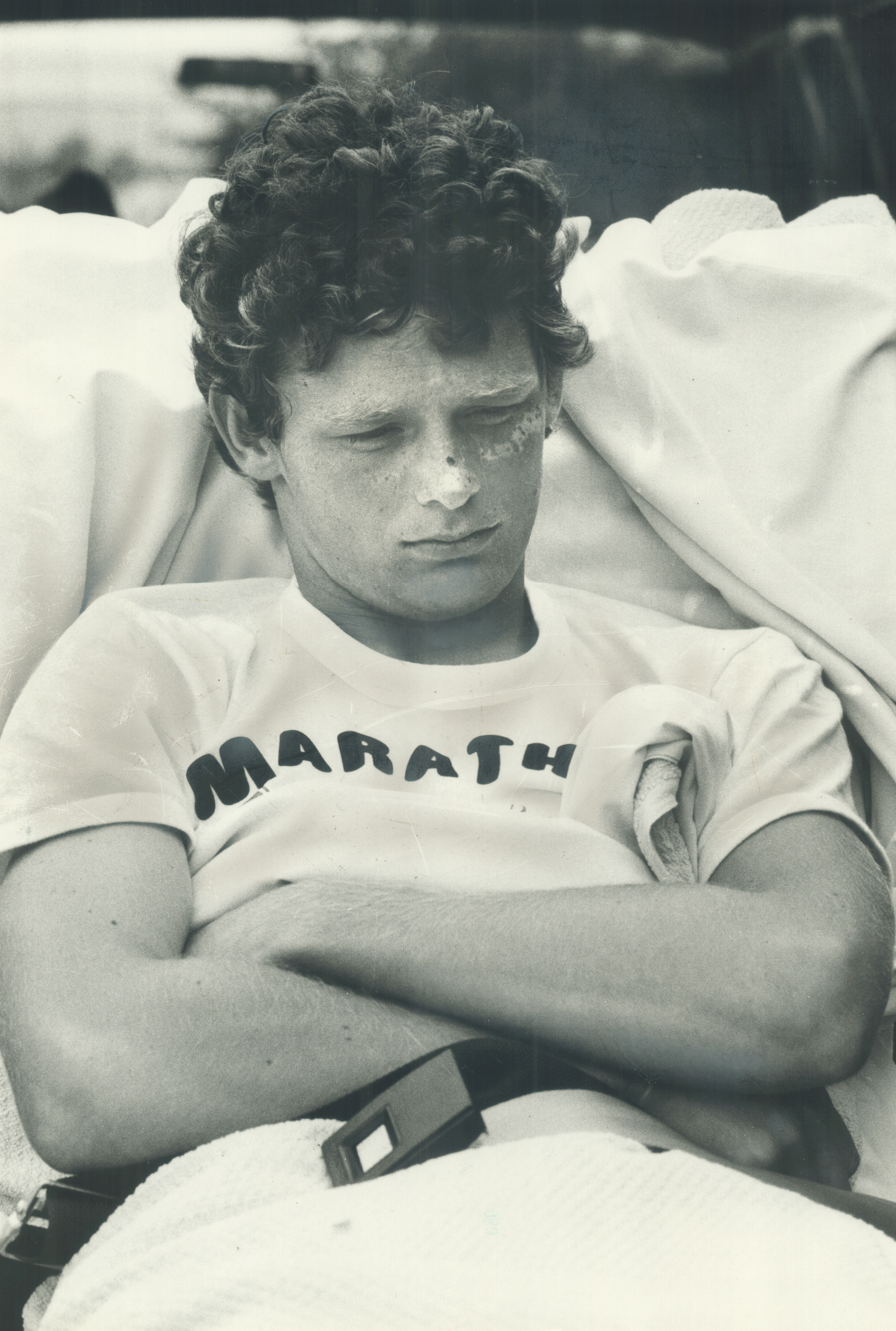 Terry Fox, lying down and surrounded by microphones, speaks to reporters during an event. People around him are holding out microphones towards him