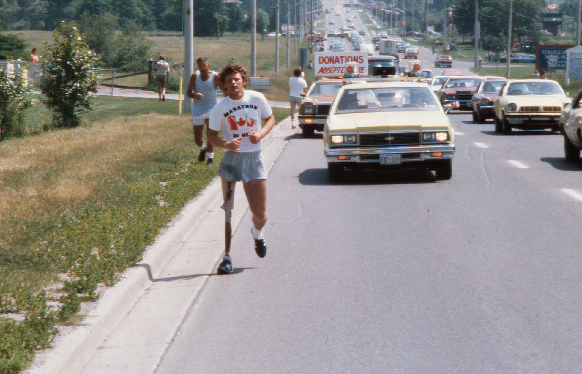 A man with a prosthetic leg, wearing a t-shirt and shorts, leads a marathon run on a busy road, with supporters and cars following behind