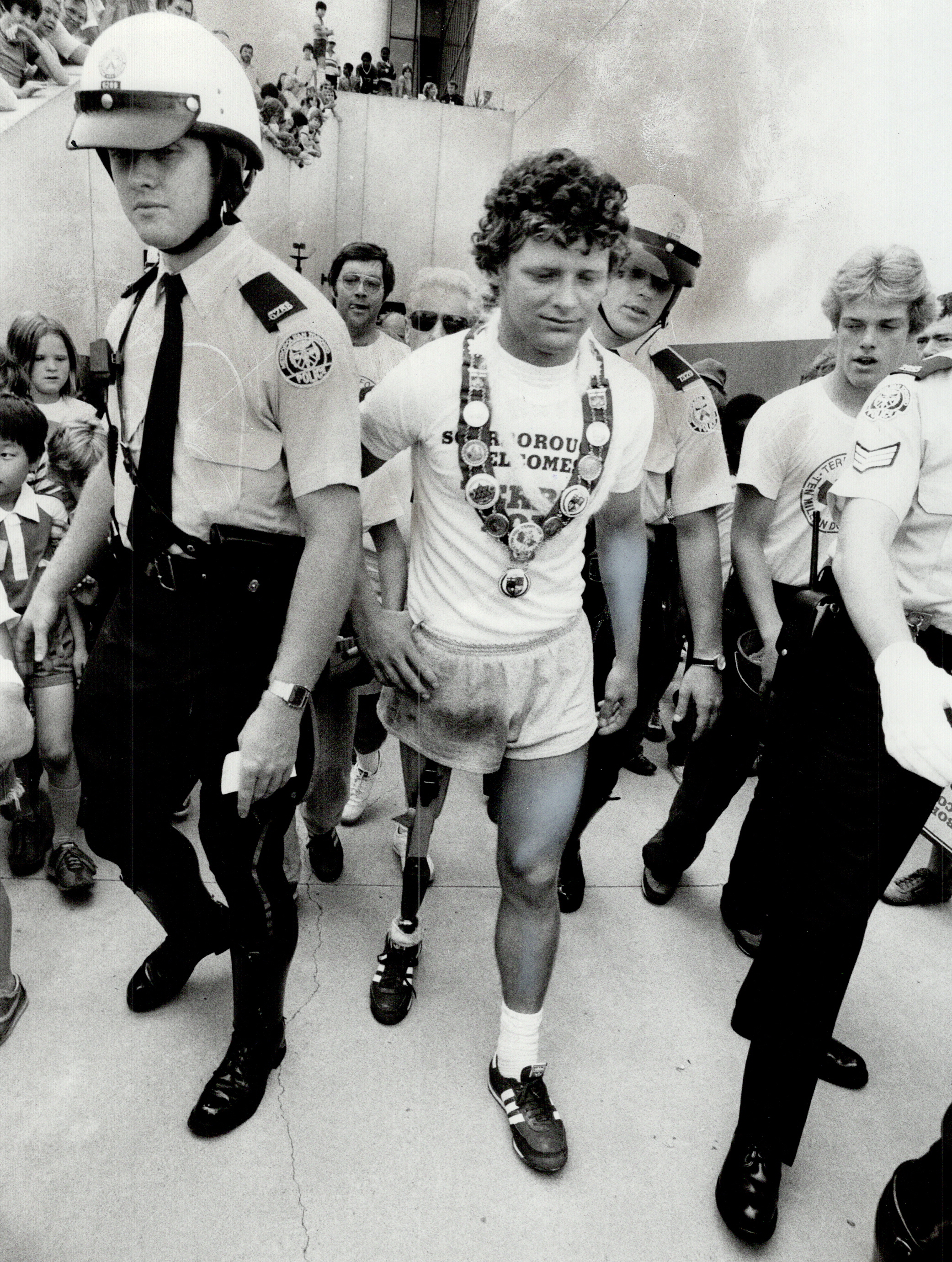 Terry Fox, wearing a prosthetic leg and sports attire, is accompanied by police officers and supporters during a public event