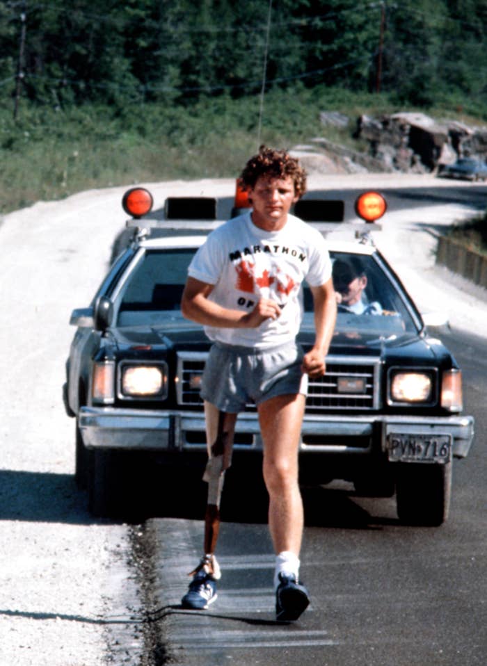 Terry Fox is running with a prosthetic leg on a road, followed closely by a support vehicle in the background. He is wearing a "Marathon of Hope" T-shirt and shorts