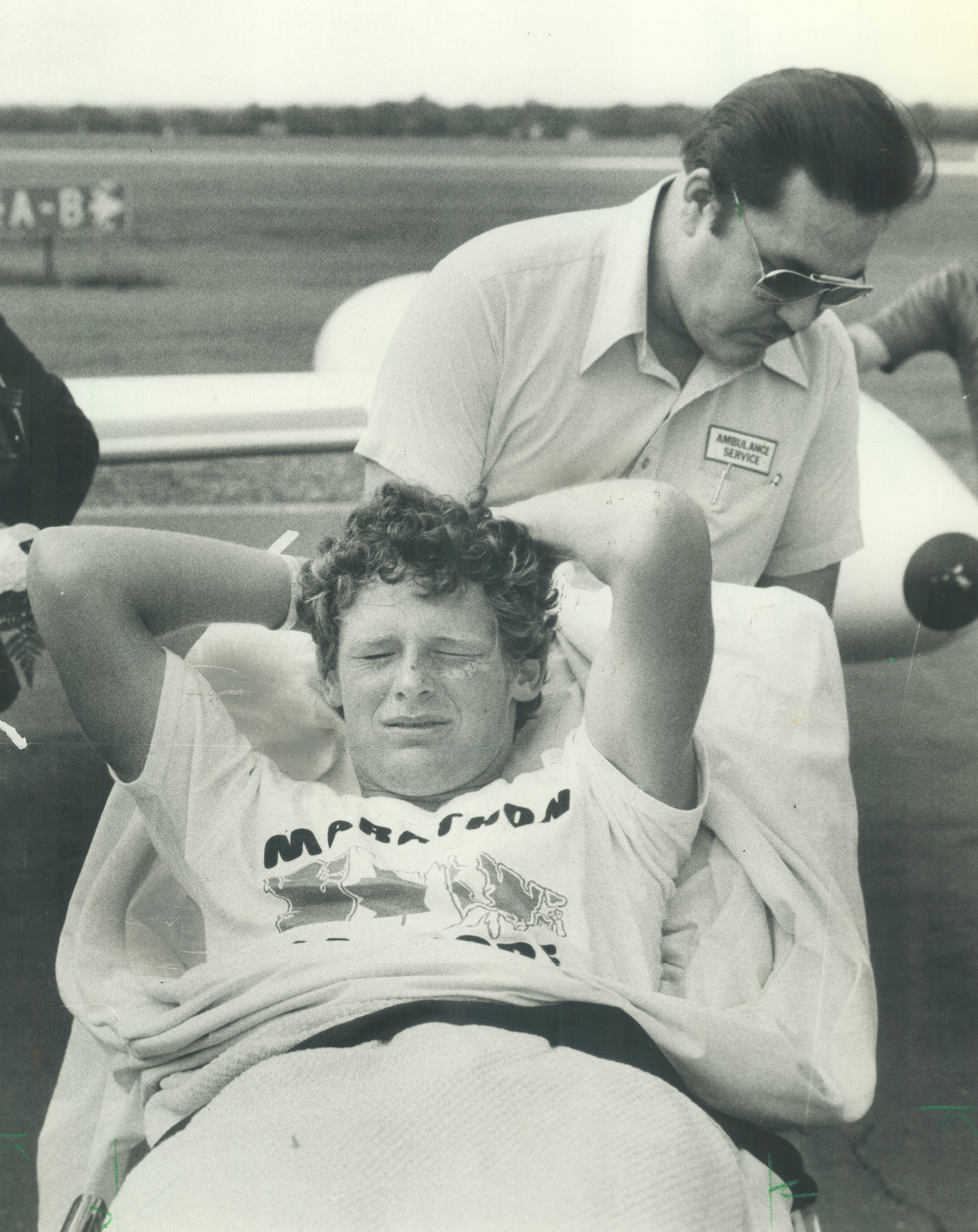 Terry Fox, wearing a marathon t-shirt, lies back with his hands behind his head while an unidentified man in a shirt and glasses assists him