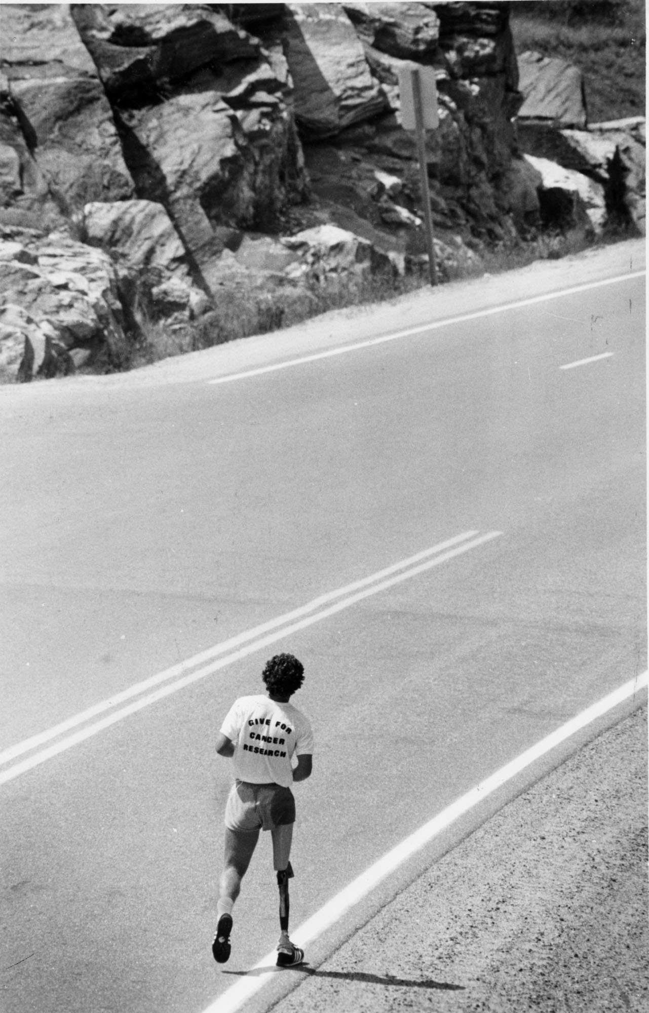 Person with a single leg prosthesis running down a highway with rocky terrain in the background. The back of their shirt reads "Marathon of Hope - 1980."
