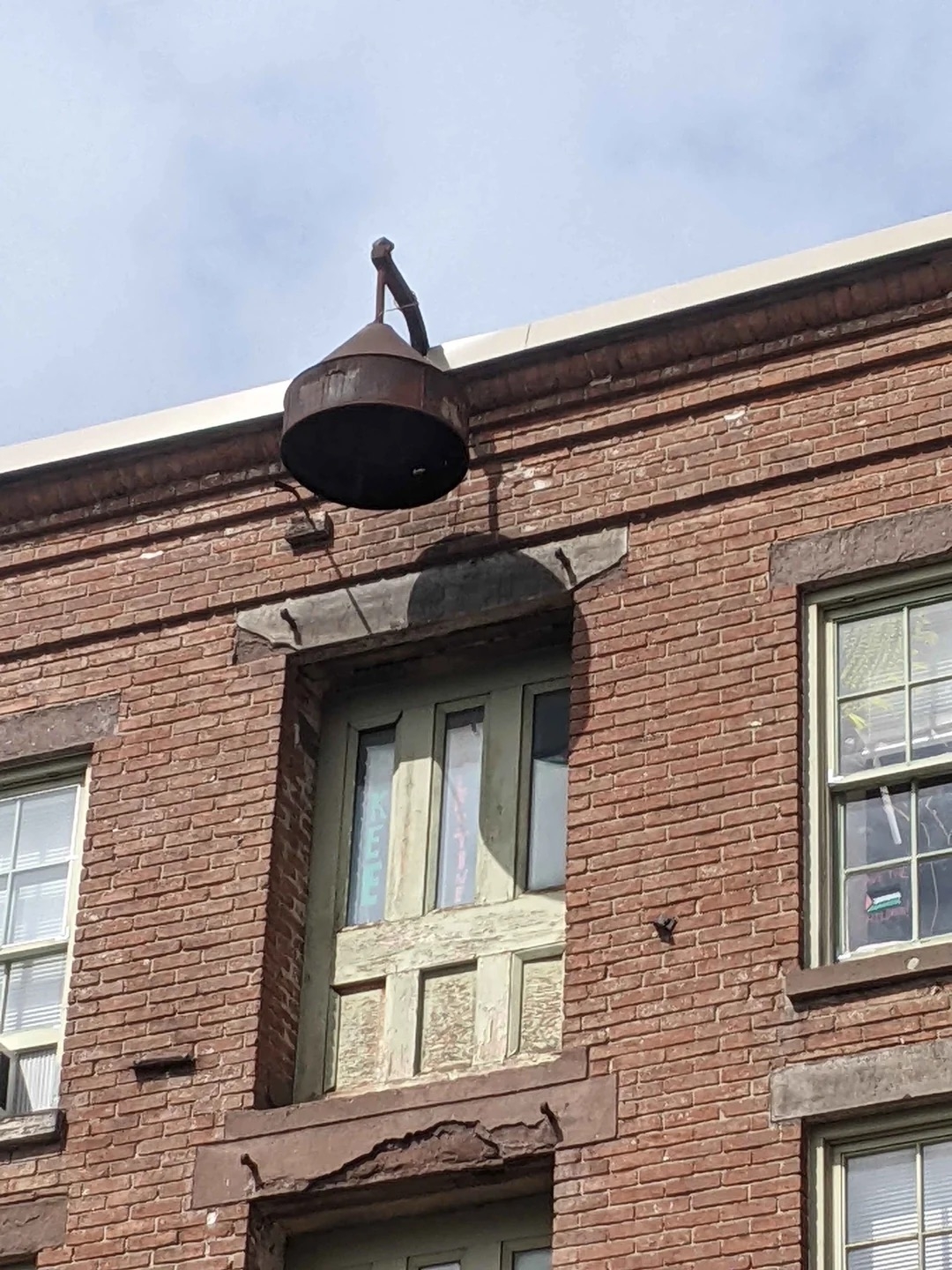 Old, aged brick building with a rusted fire escape door and a large metal light fixture-shaped item above it. Some windows are visible on either side