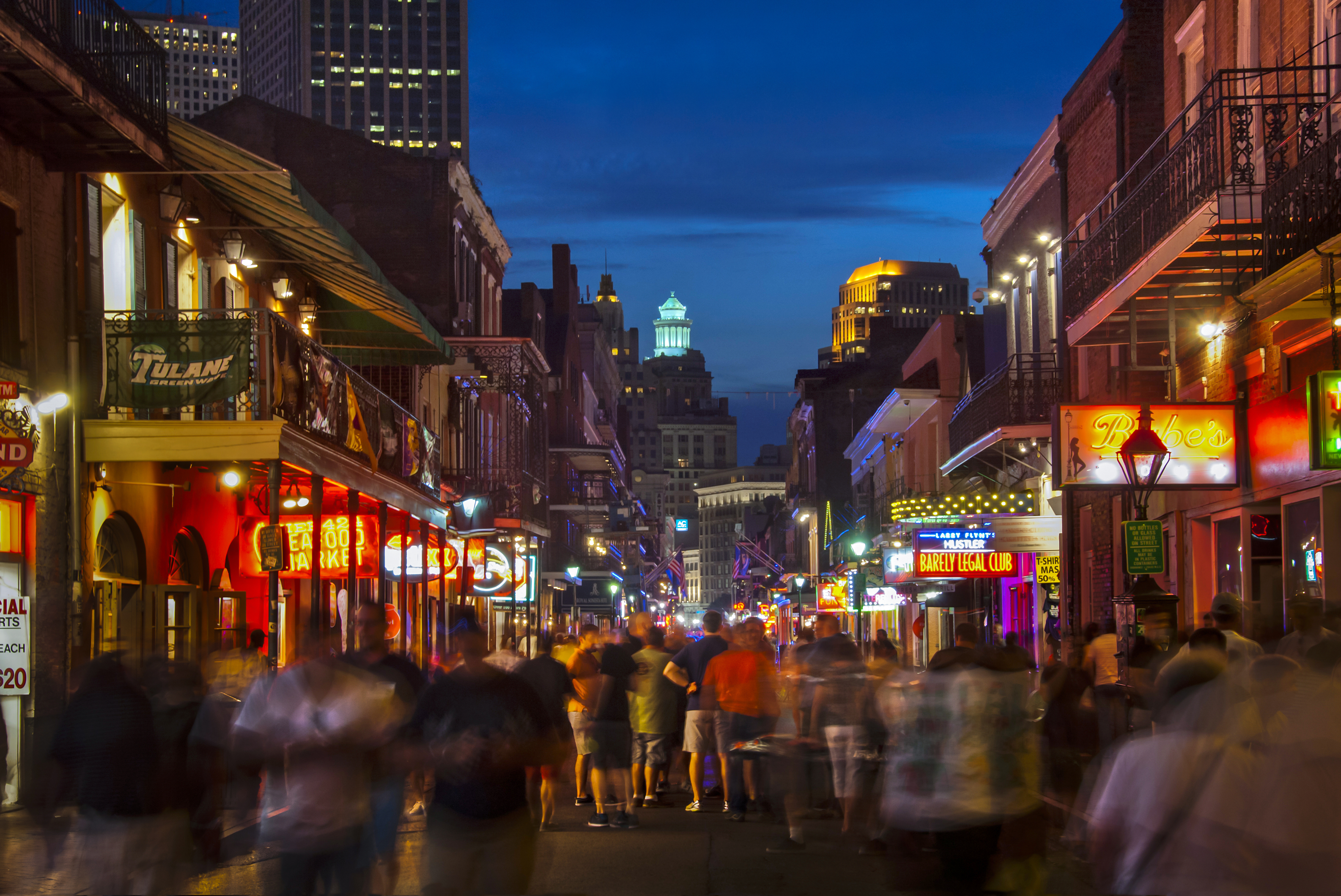 A lively, bustling street scene in a city at night, filled with people walking past brightly lit shops, bars, and restaurants