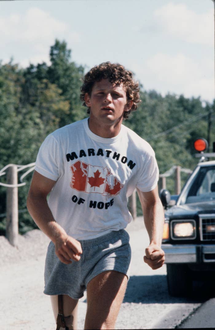 Terry Fox, wearing a "Marathon of Hope" shirt, runs on a gravel road with a vehicle trailing behind him