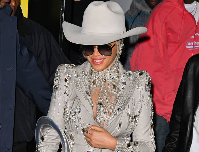Beyoncé in a rhinestone-embellished suit, large hat, and sunglasses, smiles while attending an event