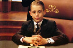 Macaulay Culkin, as Richie Rich, sits at a desk in a suit with his hands clasped, a magnifying glass in front of him