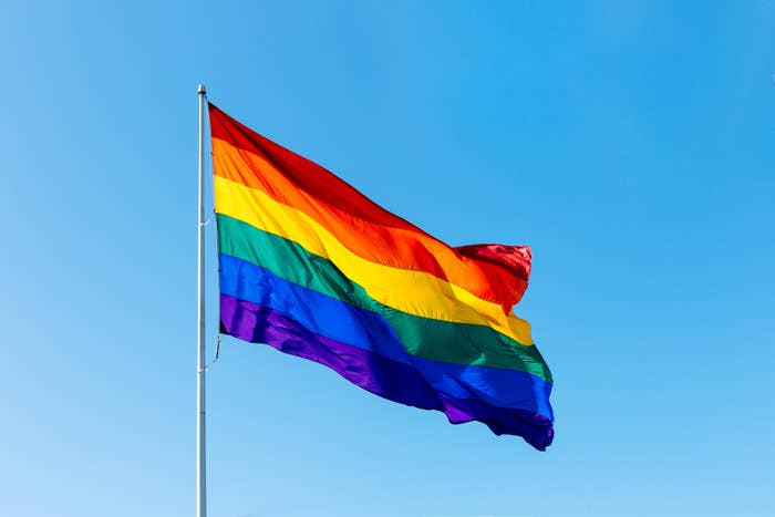 A large multi-colored rainbow flag waves against a clear blue sky, symbolizing LGBTQ+ pride