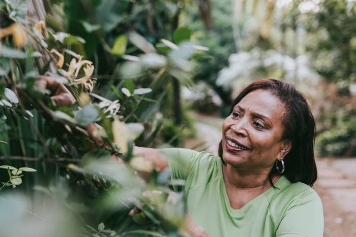 A woman smiles while gardening outdoors, surrounded by greenery