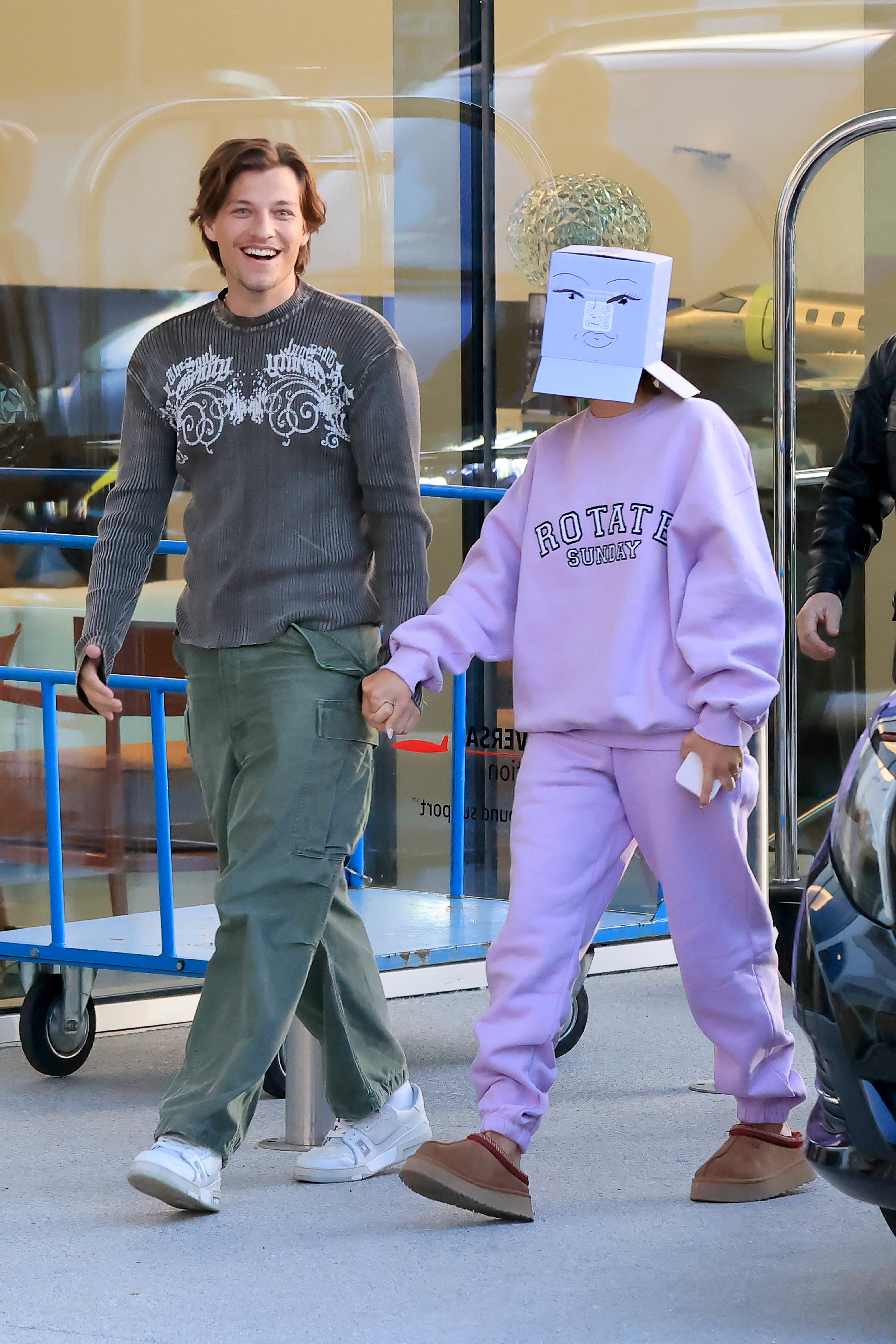 Jake and Millie in a tracksuit, boots, and a paper bag over her head with a face drawing, holding hands and walking outside