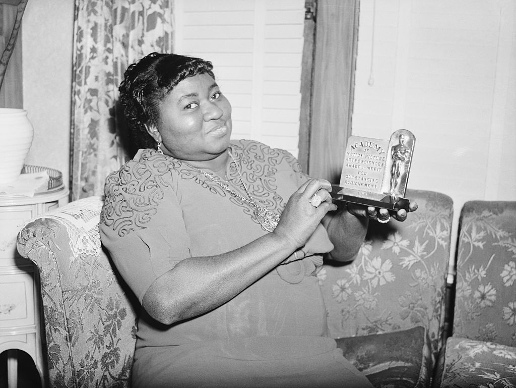 Hattie McDaniel smiling and holding an Academy Award Oscar statuette while sitting on a floral-patterned sofa