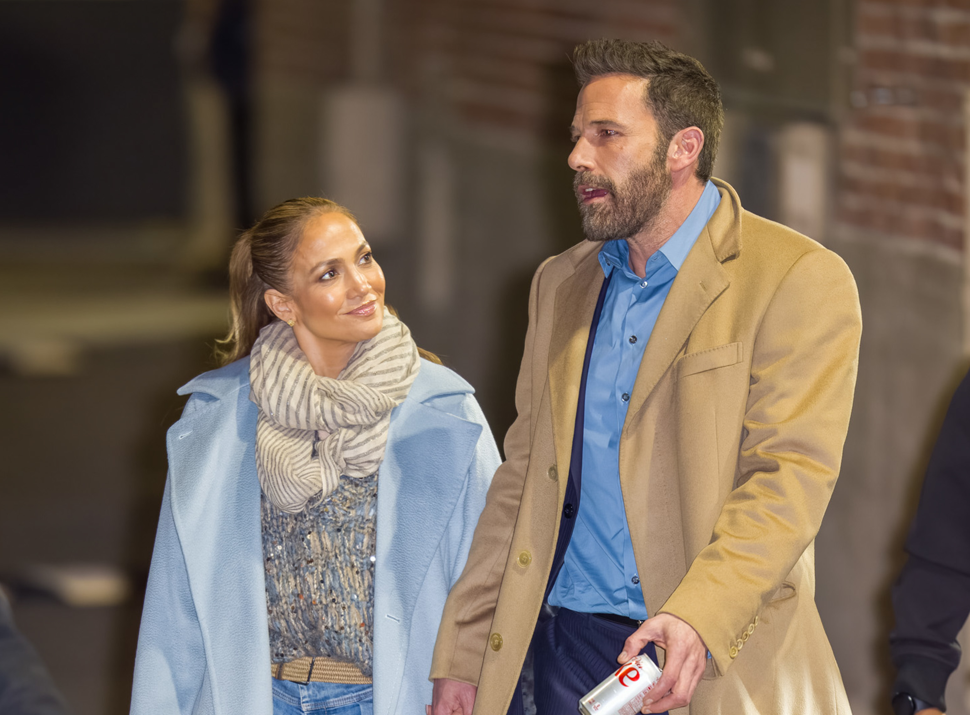 Jennifer Lopez and Ben Affleck walking together, both dressed in casual yet stylish outfits. Jennifer wears a cozy sweater and scarf, while Ben sports a coat over a shirt
