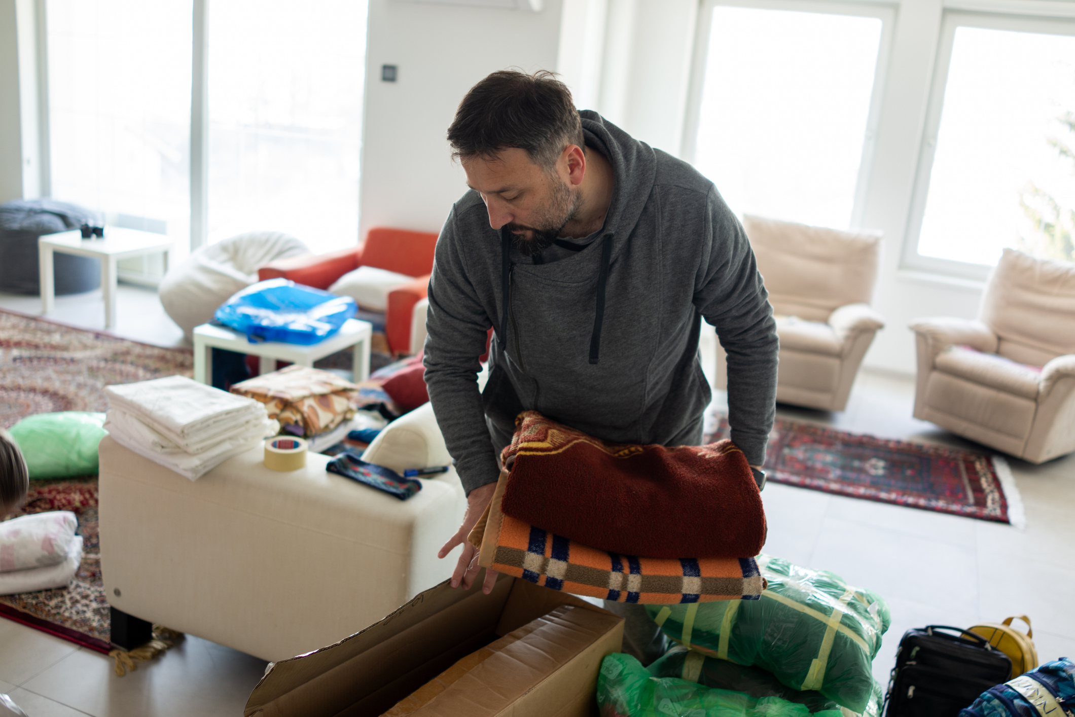 A man is shown in a living room, organizing and packing blankets into a cardboard box surrounded by various household items