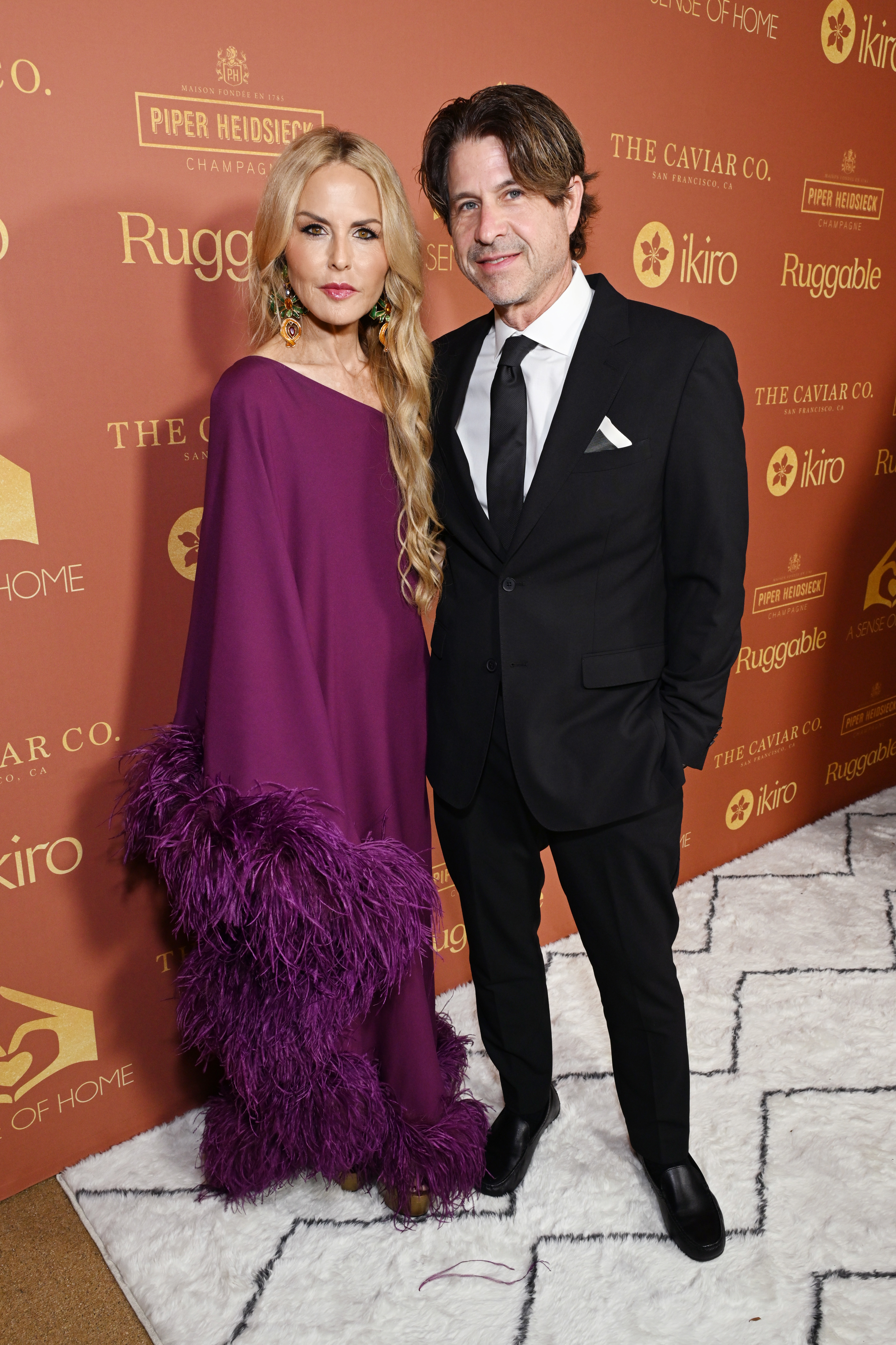 Rachel Zoe, in a flowing dress with feathered sleeves, and Rodger Berman, in a suit, pose together at a red carpet event
