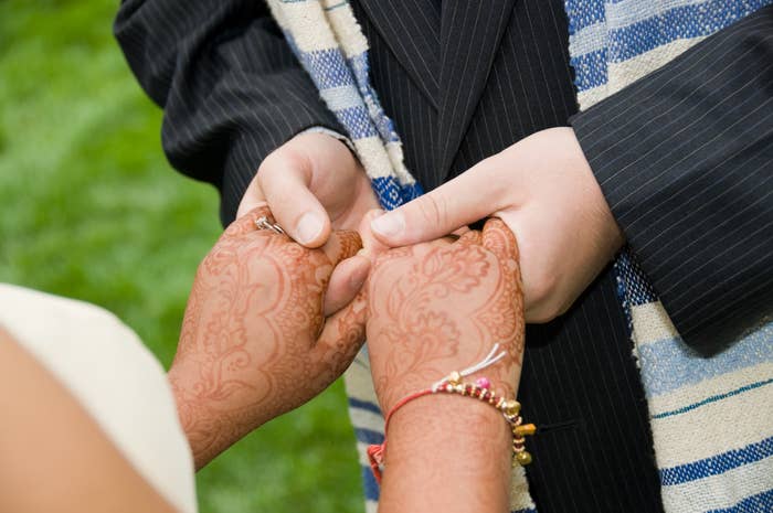 Close-up of a couple holding hands during a wedding ceremony. The bride's hands have intricate Mehndi designs, and the groom's hands are dressed in a striped suit jacket