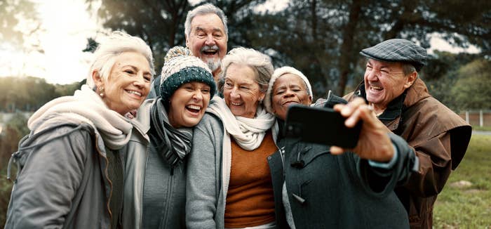 A group of six elderly people smile warmly and huddle together for a selfie outdoors, some wearing winter hats and jackets