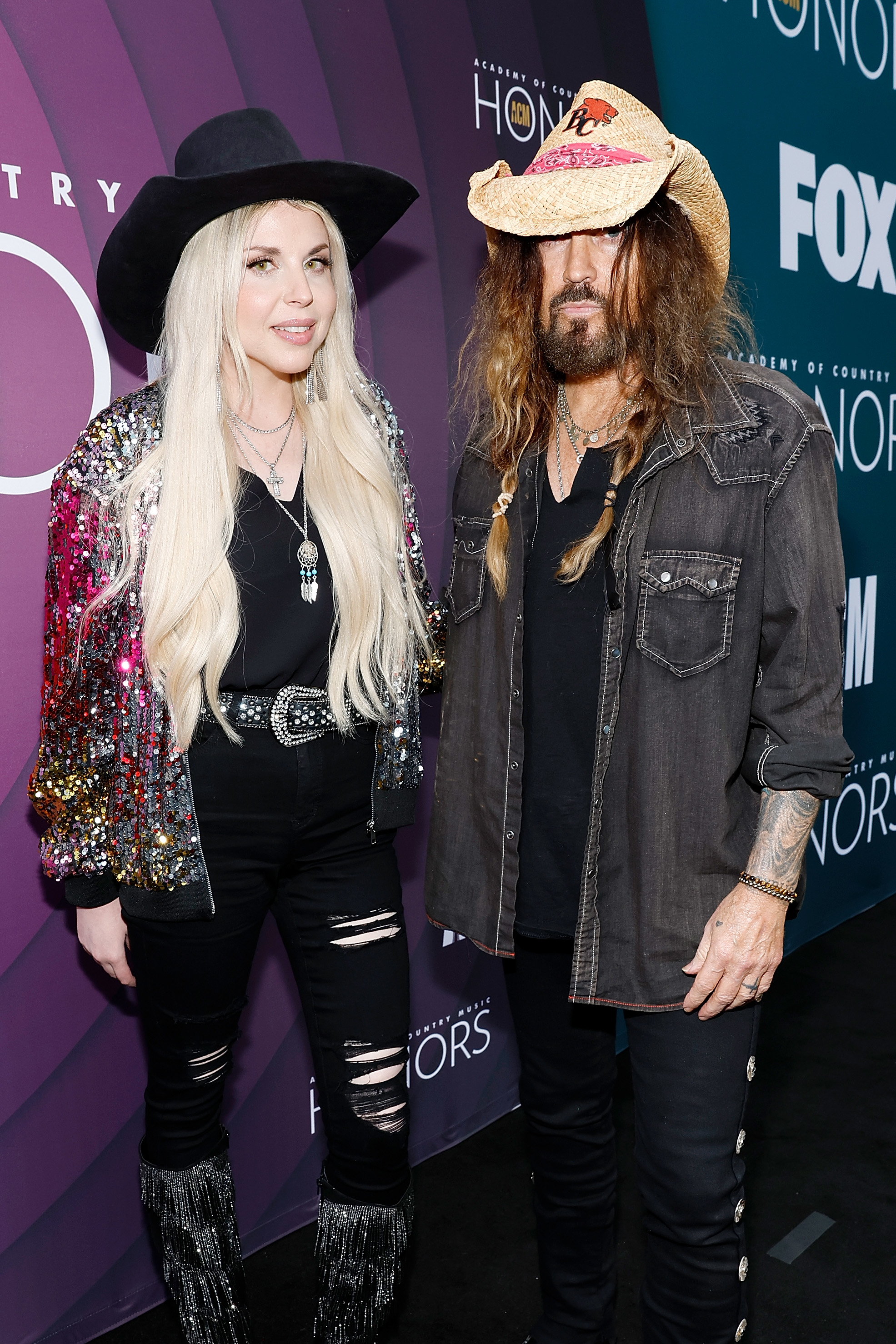 Billy Ray Cyrus and Firerose at a red carpet event: Billy Ray in a cowboy hat, dark button-down shirt, and jeans; Firerose in a sequin jacket, hat, and jeans