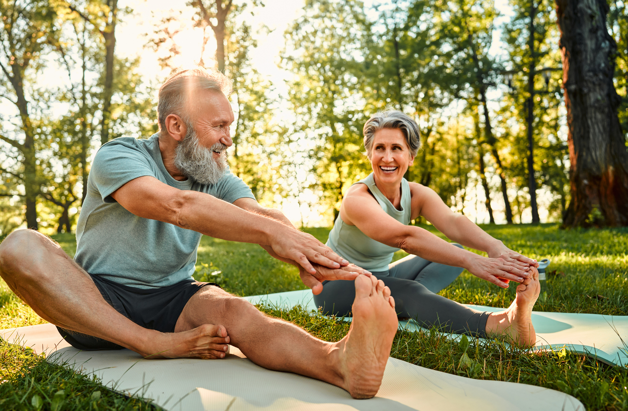 An elderly man and woman, smiling, sit on yoga mats in a park, stretching and touching their feet