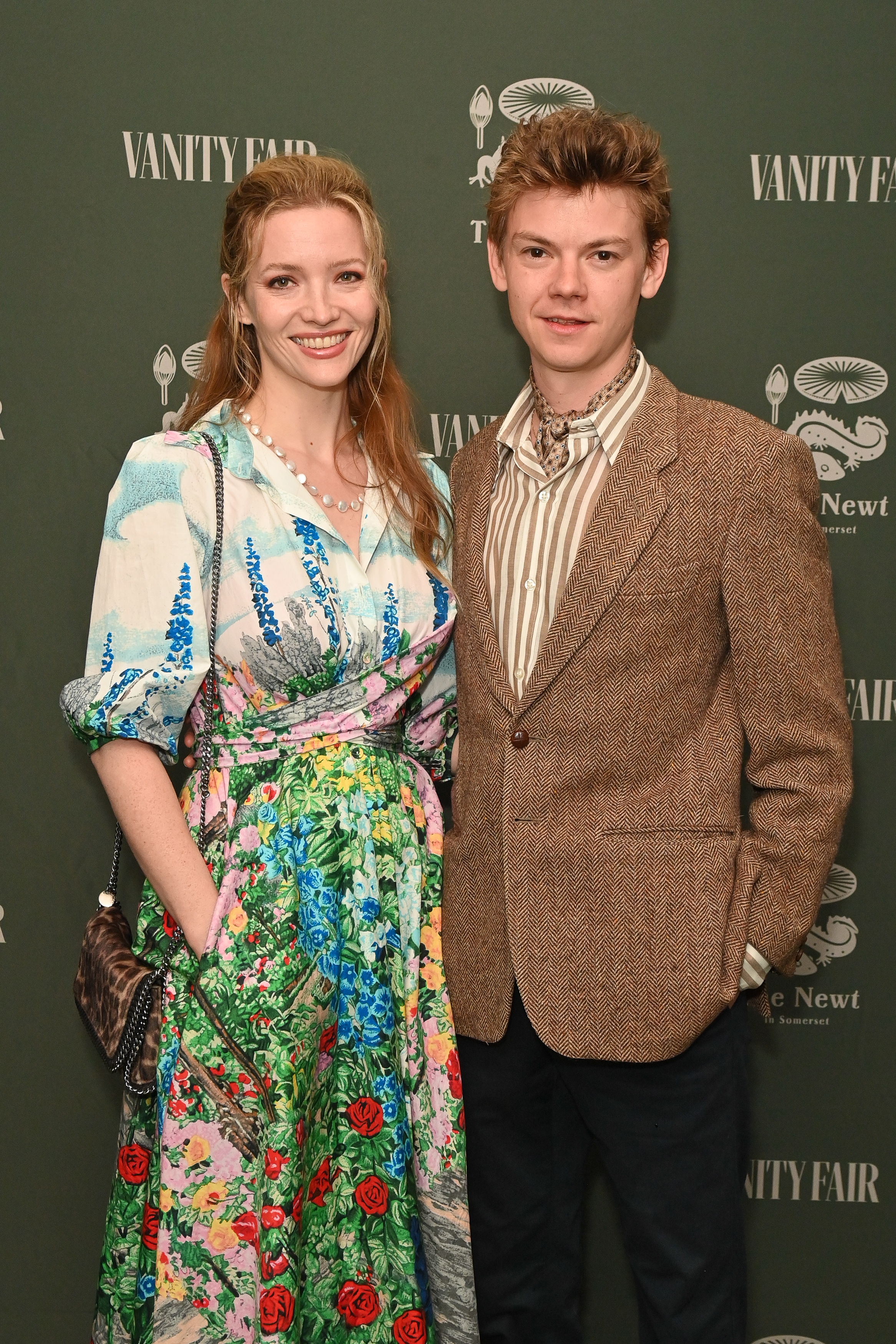 Talulah Riley in a floral dress and Thomas Brodie-Sangster in a striped shirt with a blazer pose together at the Vanity Fair event