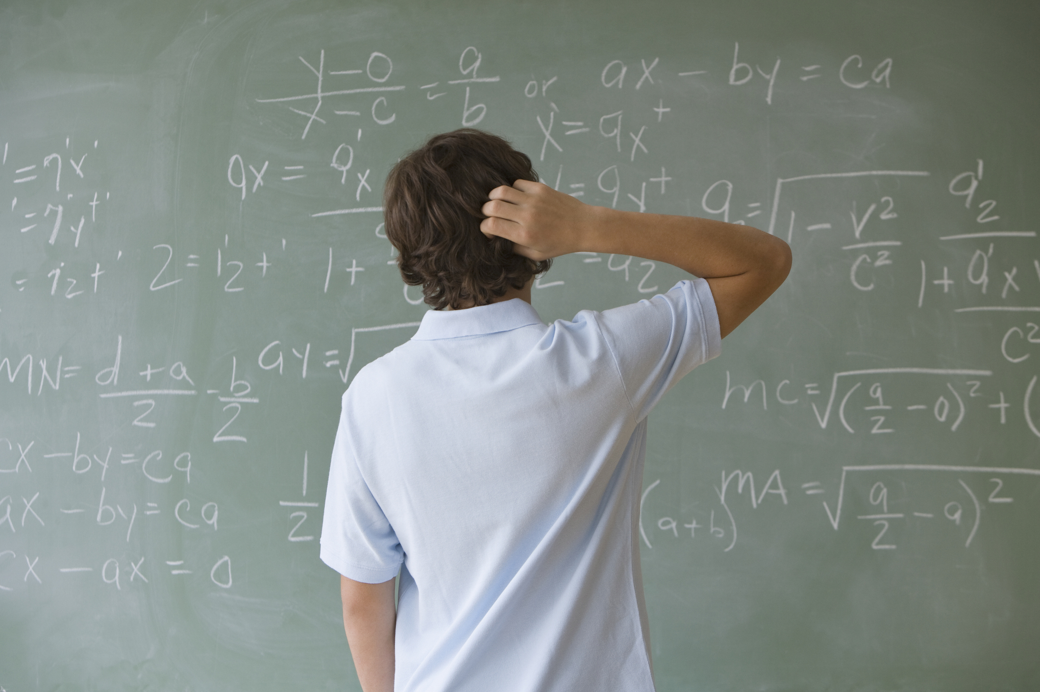 A person with short hair, wearing a light-colored shirt, stands in front of a chalkboard filled with complex math equations, scratching their head in confusion