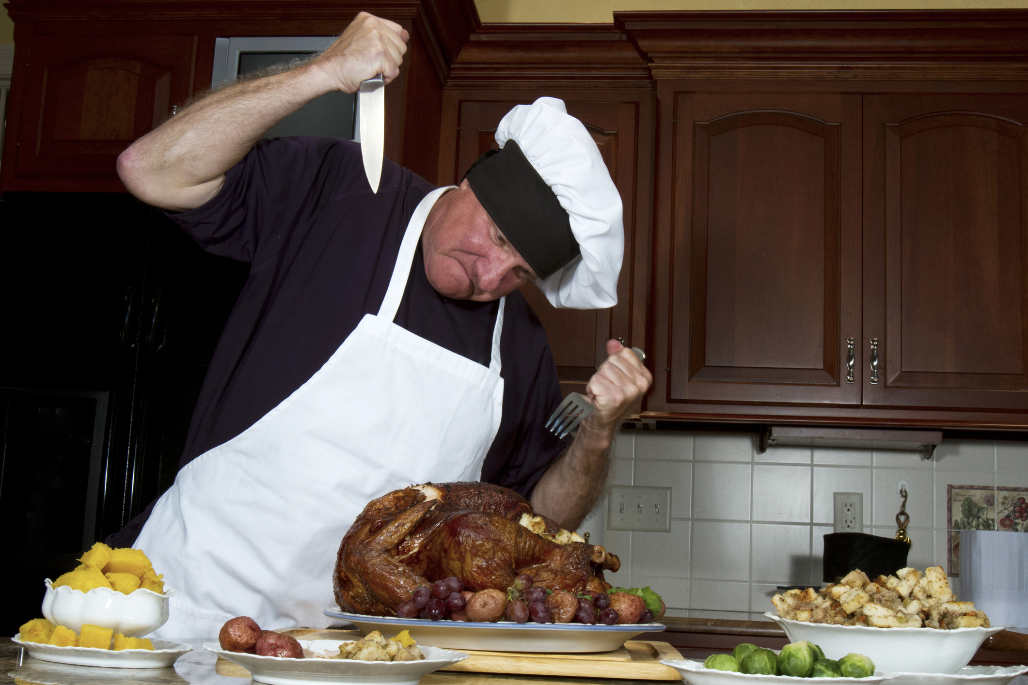 A man dressed as a chef dramatically holds a knife and fork above a large roasted turkey in a kitchen, ready to carve it. Various side dishes surround the turkey